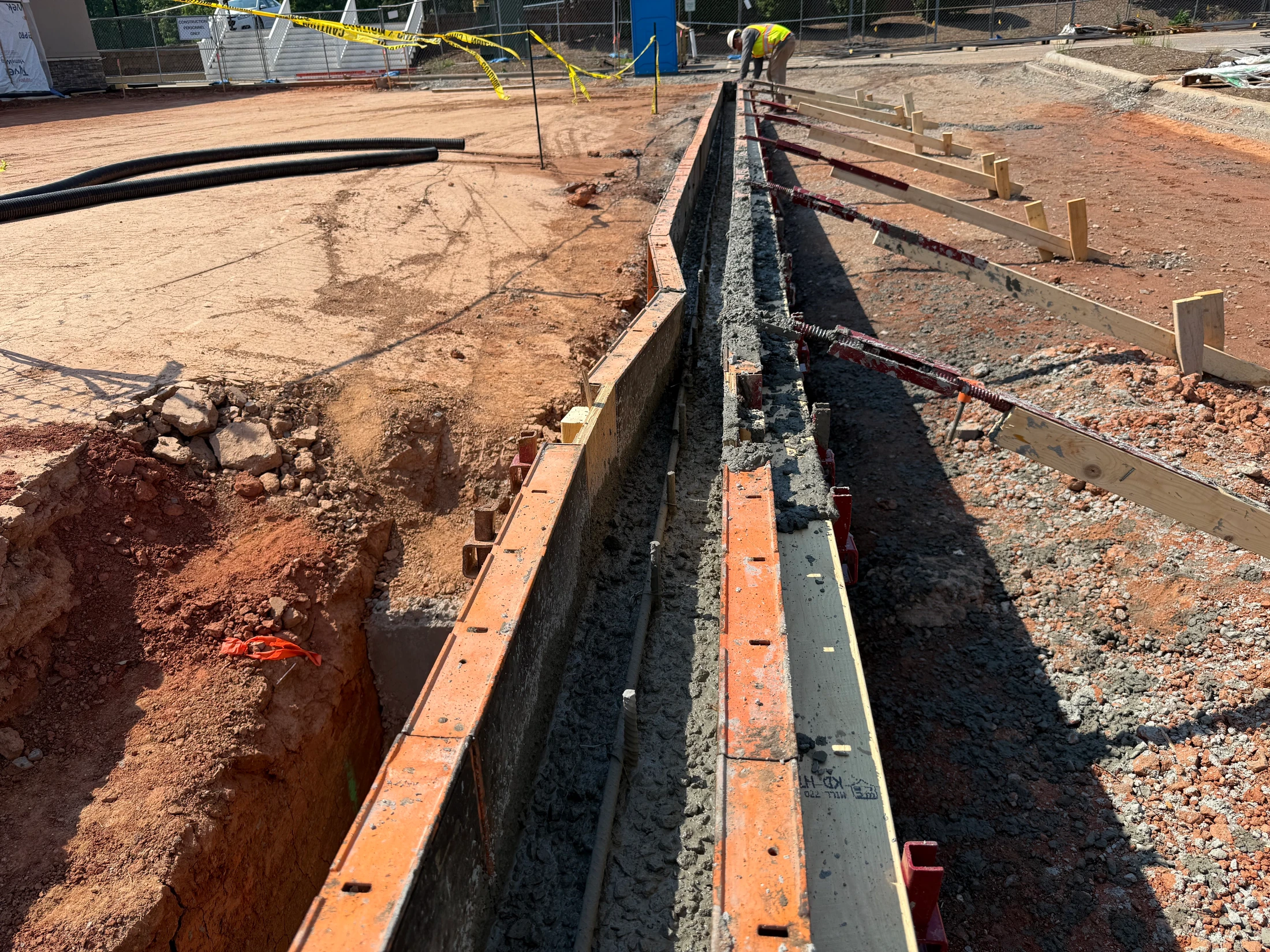 A long formwork structure filled with wet concrete for a foundation on a construction site, with a worker in the background adjusting supports.