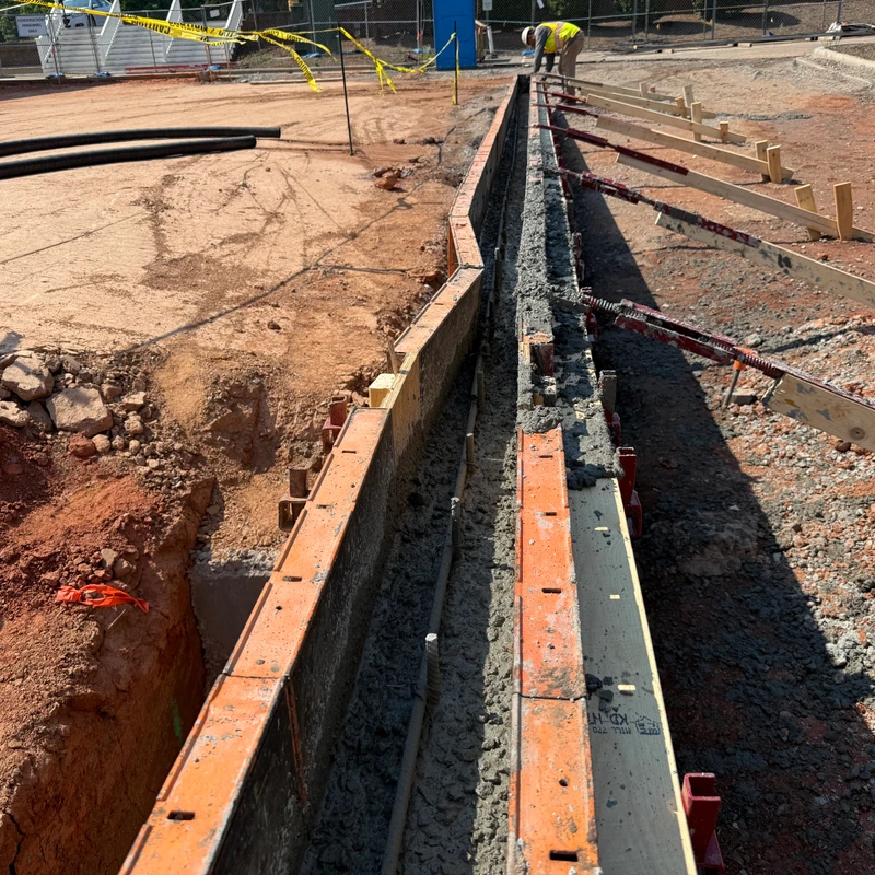 A long formwork structure filled with wet concrete for a foundation on a construction site, with a worker in the background adjusting supports.