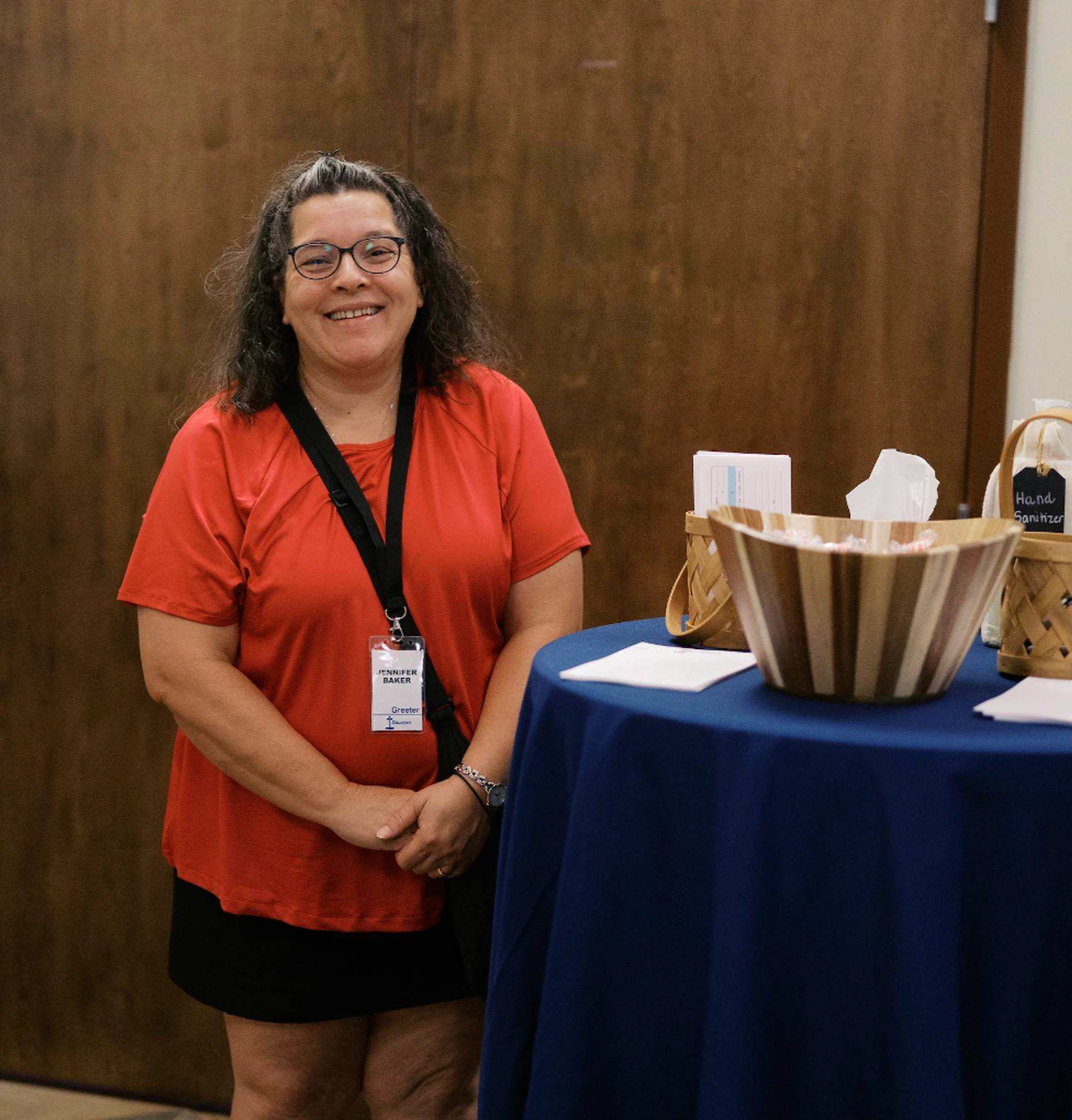 A smiling woman with glasses and a red shirt stands next to a welcome table. She wears a name tag that identifies her as Jennifer Baker, a greeter.