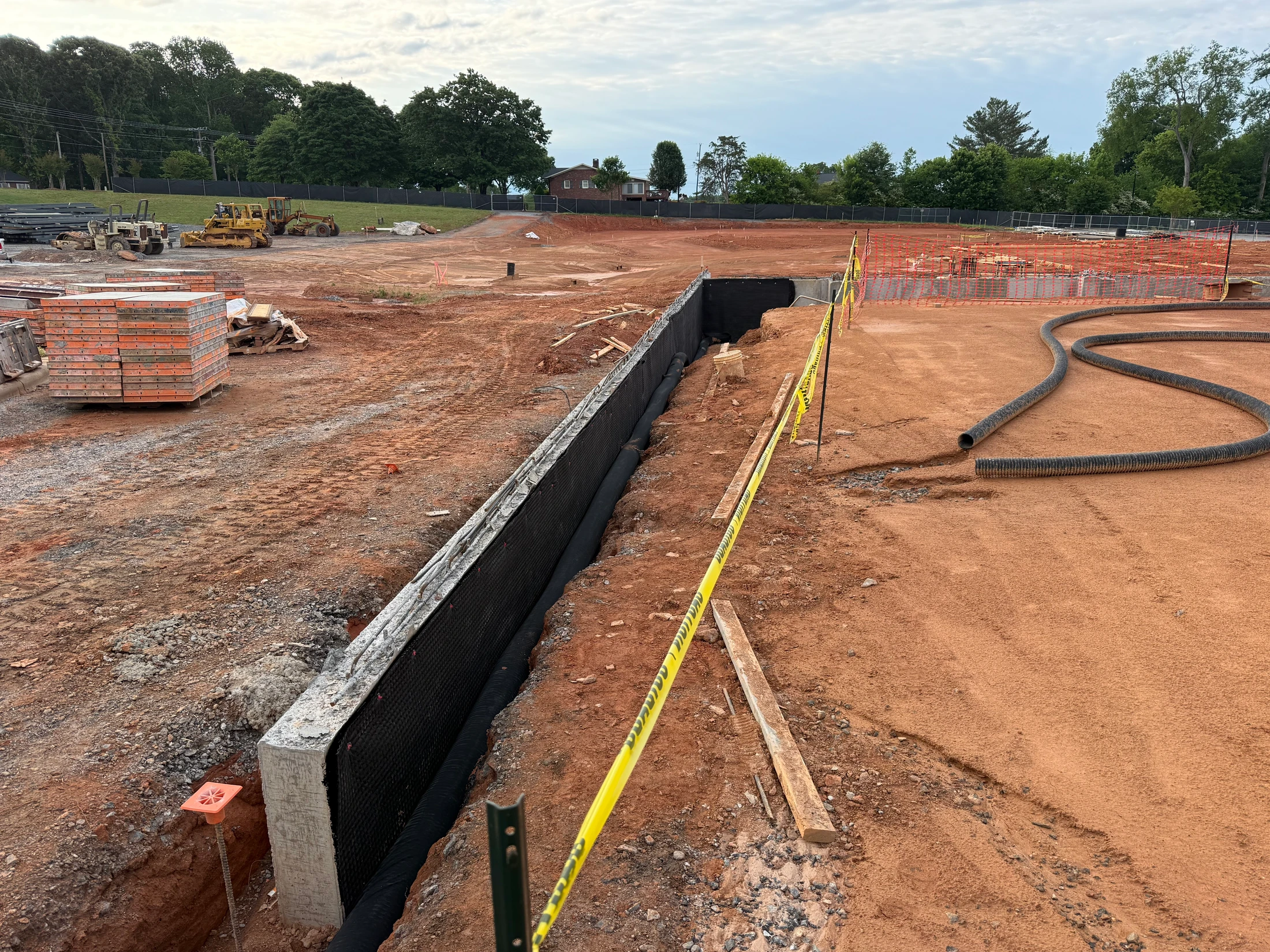 A wide view of a construction site with red dirt. In the foreground, a concrete foundation wall is being waterproofed, sectioned off by yellow caution tape.