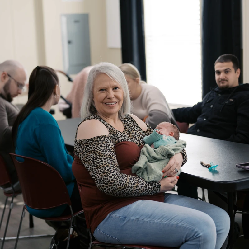 A woman with white hair sits at a table, smiling at the camera as she cradles a newborn baby. Other people are seated in the background at the same table.