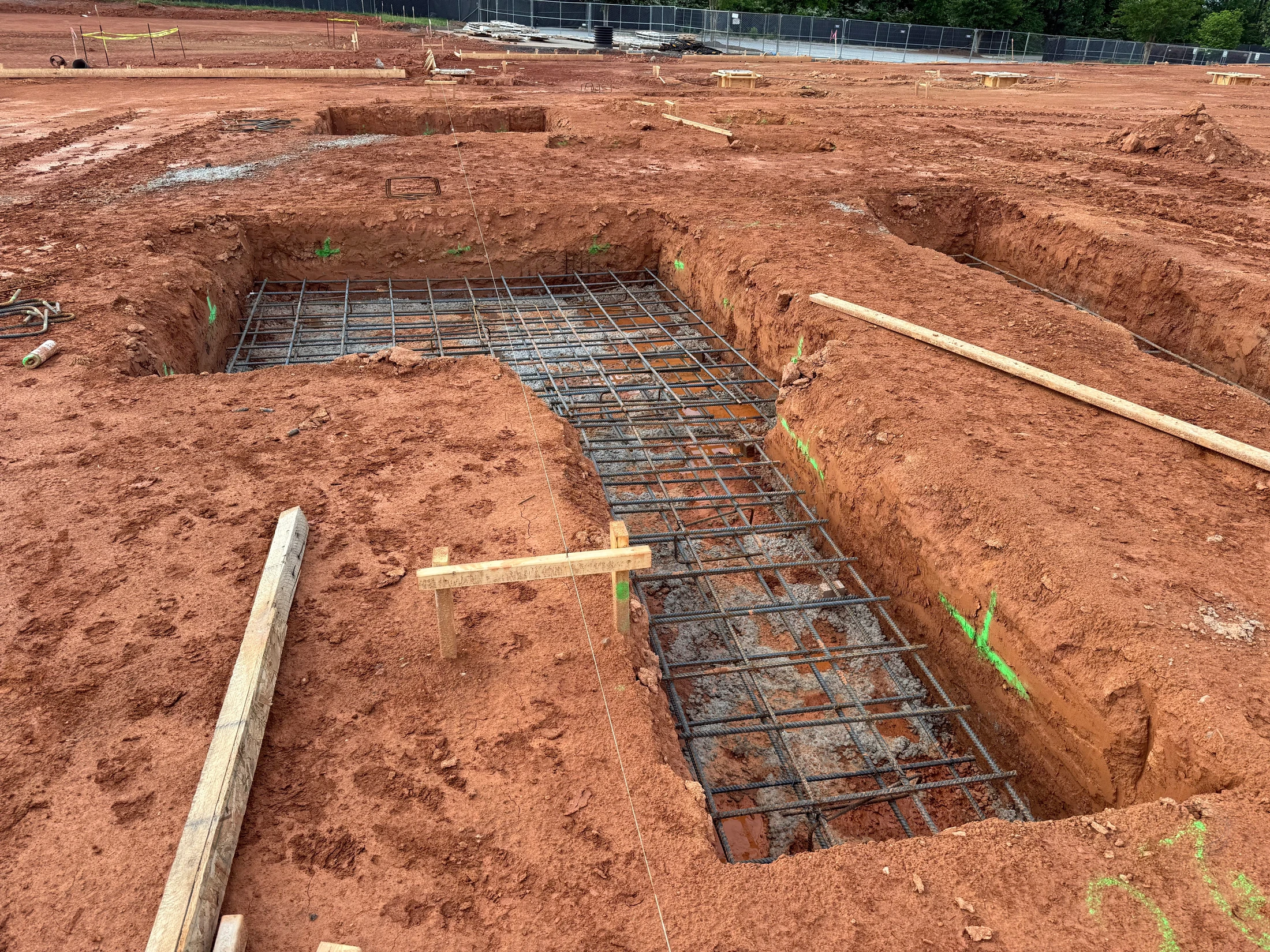 A construction site with trenches dug into red clay soil. A steel rebar grid is laid inside a trench, preparing for a concrete foundation to be poured.