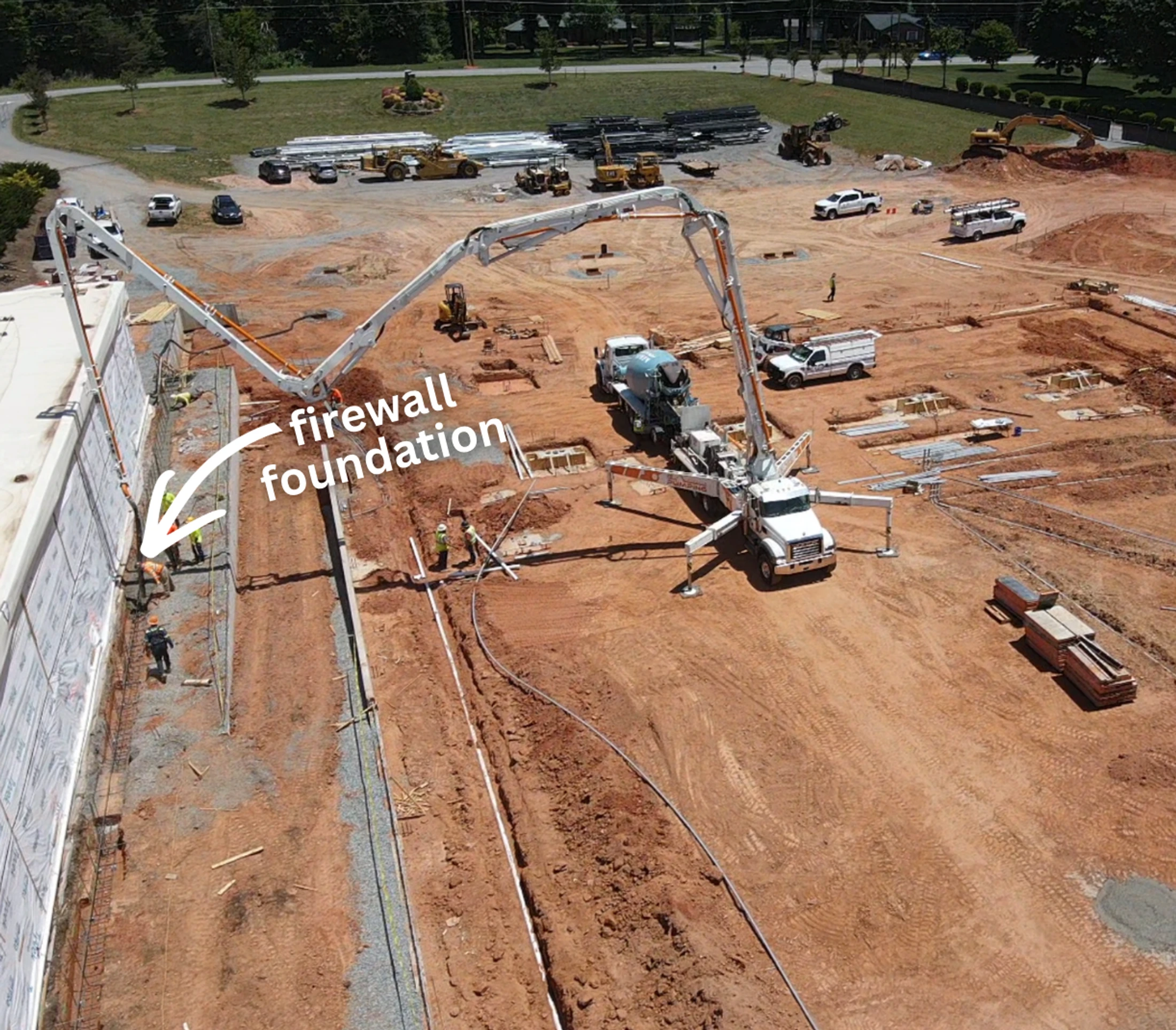 Aerial view of a construction site where a large concrete pump truck is pouring a new firewall foundation next to an existing building, with workers nearby.