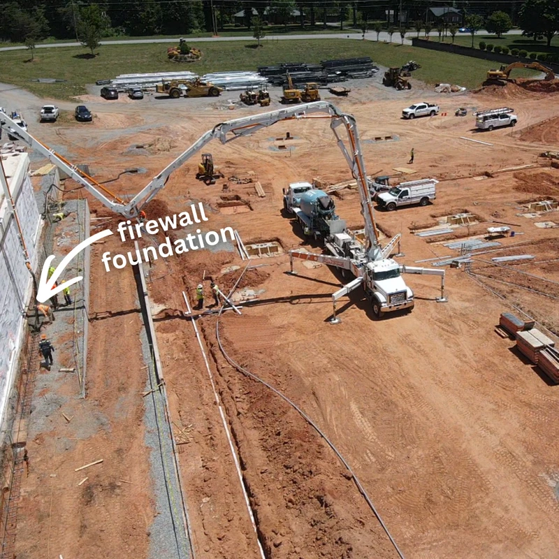 Aerial view of a construction site where a large concrete pump truck is pouring a new firewall foundation next to an existing building, with workers nearby.