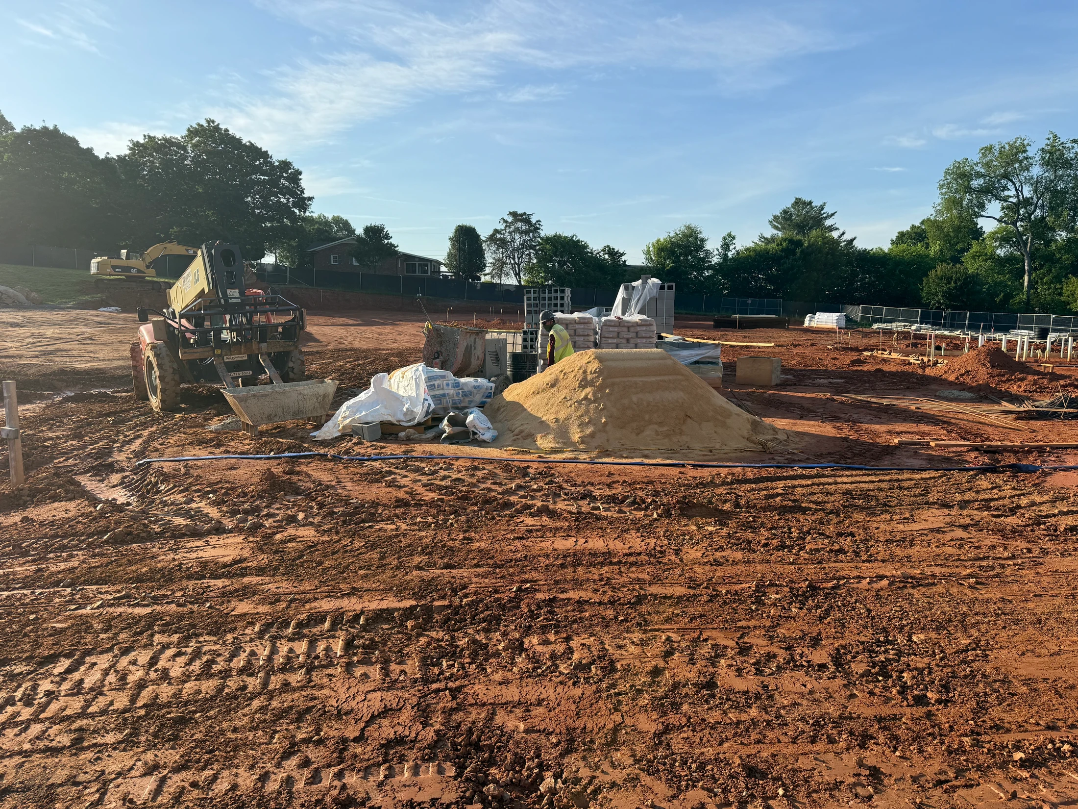 A construction site with red clay ground, a pile of sand, building materials, and a forklift under a clear blue sky with some trees in the background.