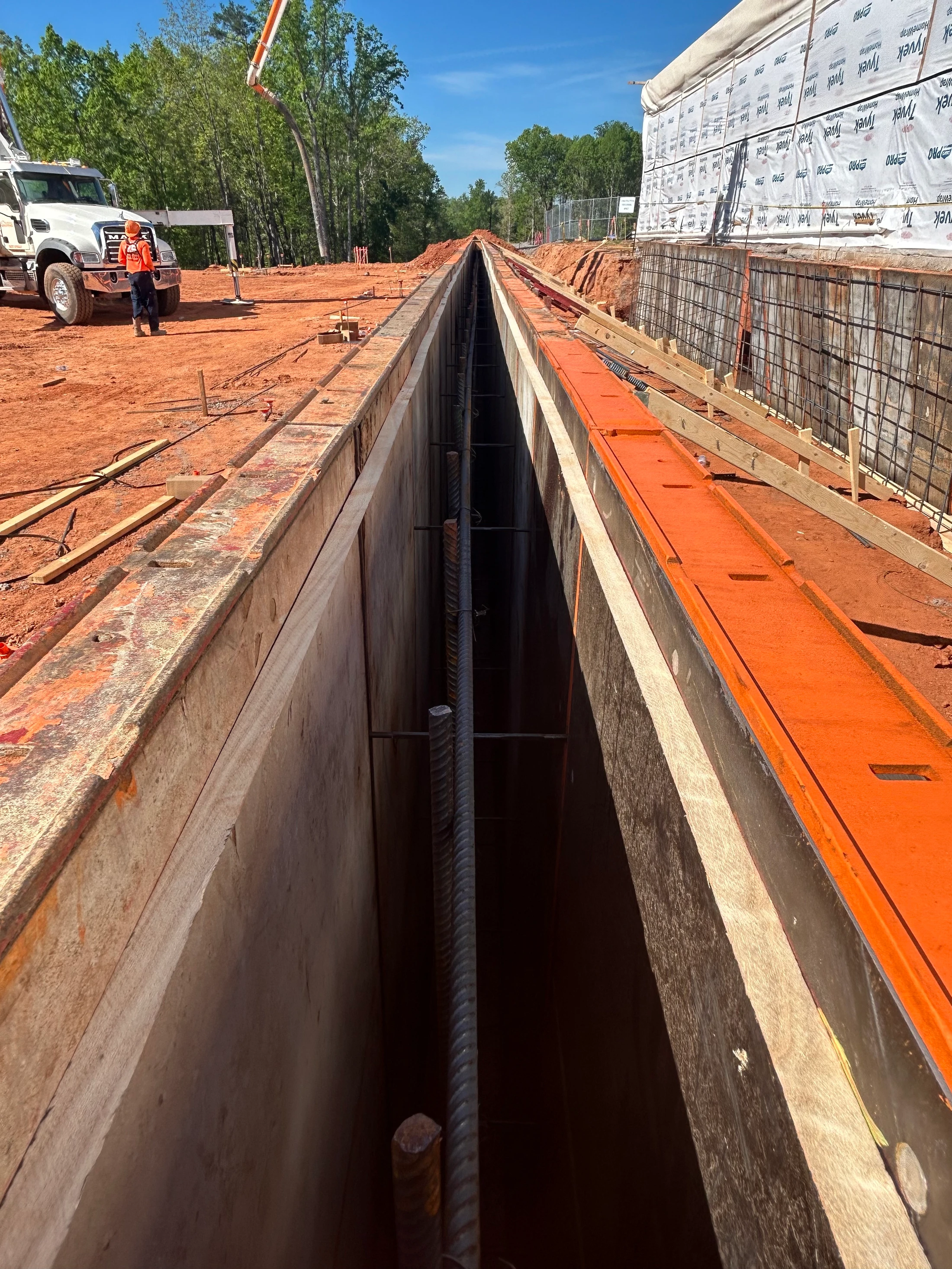 A view looking down a long, deep trench lined with concrete forms and rebar, with a worker and a concrete pump truck in the background.
