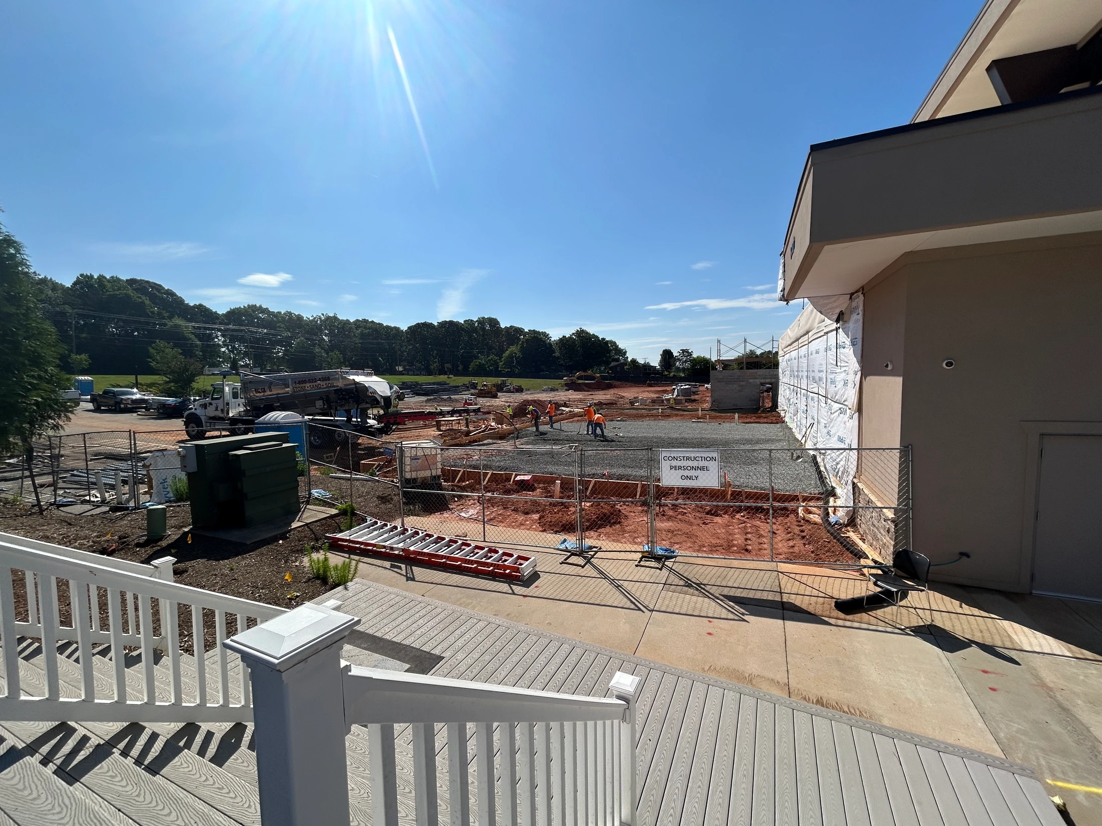 High-angle view from a wooden deck overlooking an active construction site. Workers are laying a foundation under a bright, sunny blue sky.