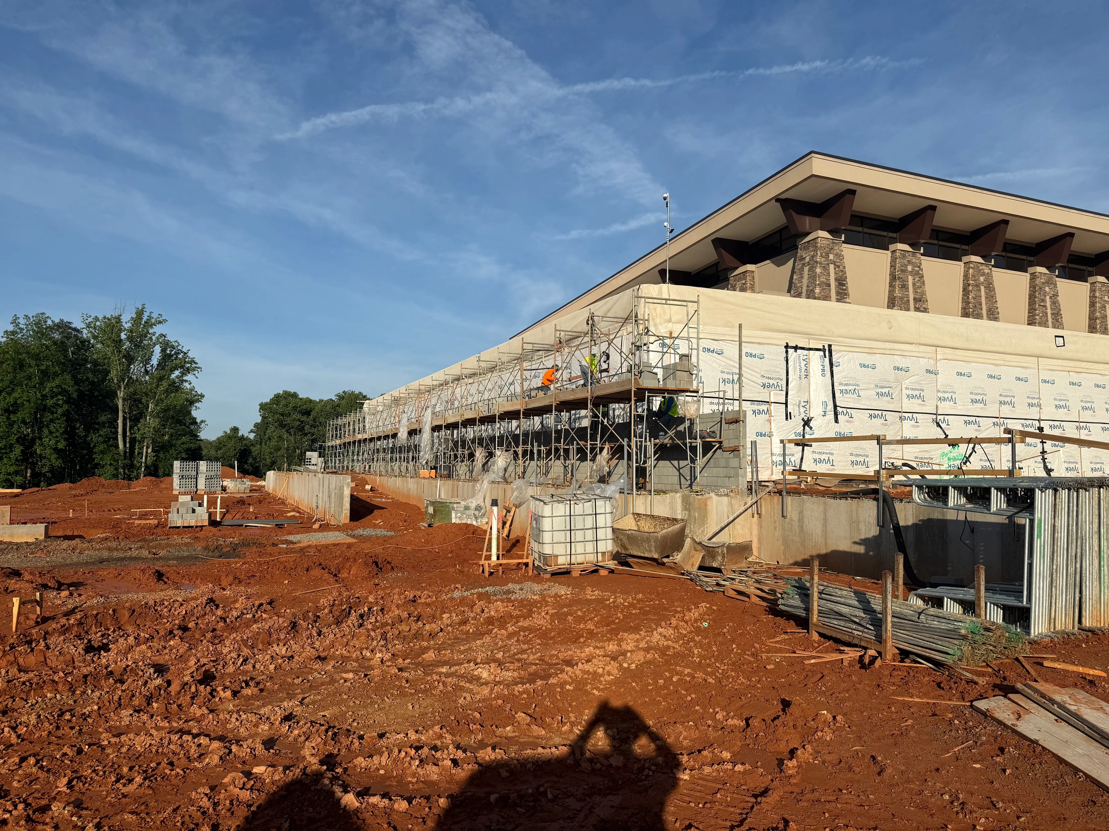 A large building under construction with extensive scaffolding along its side. Workers are visible on the scaffolding. The foreground is uneven red clay dirt.
