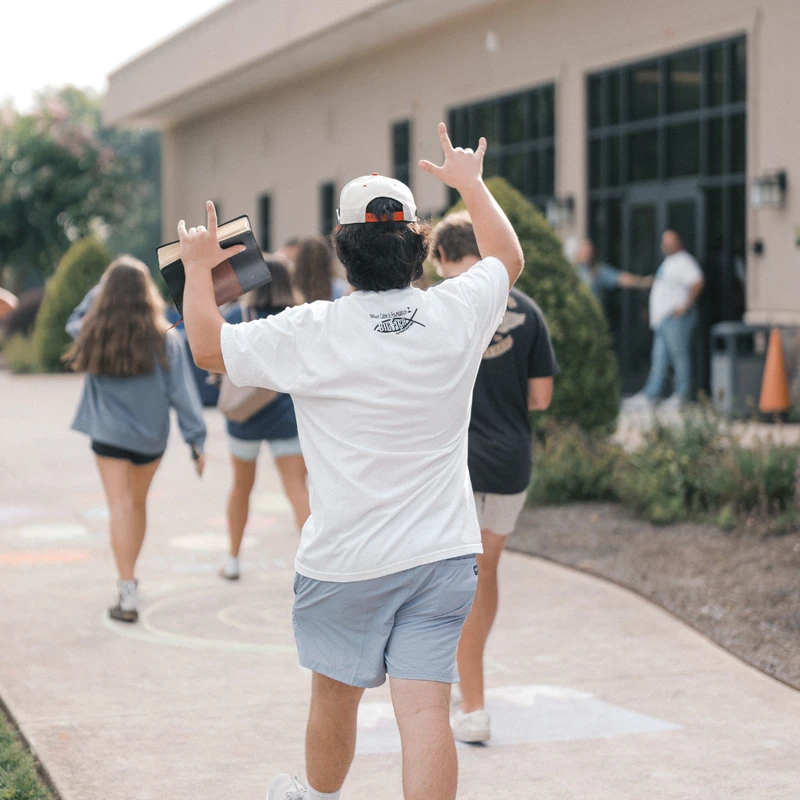 A young man, seen from behind, holds a Bible and makes a hand gesture while walking with other youths towards a building, wearing a white t-shirt and a cap.