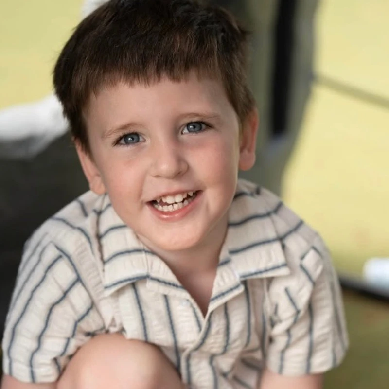 A close-up portrait of a young boy with short brown hair and bright blue eyes, smiling warmly at the camera while wearing a striped, short-sleeved shirt.
