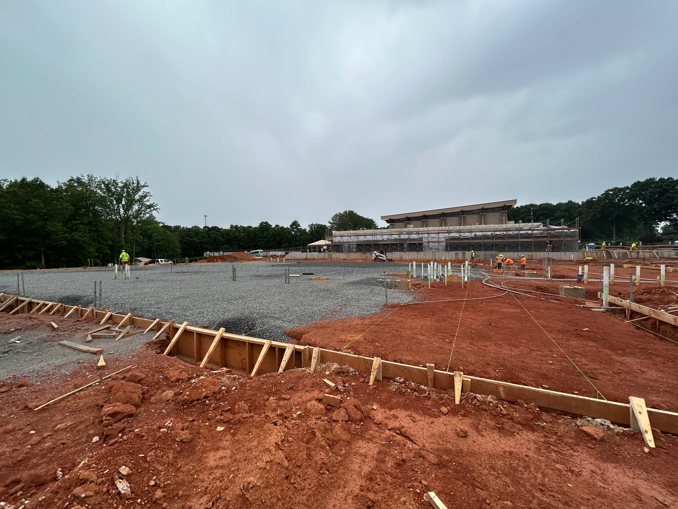 A wide view of a construction site with red clay earth, wooden formwork, and a large gravel area prepared for a foundation under an overcast sky.