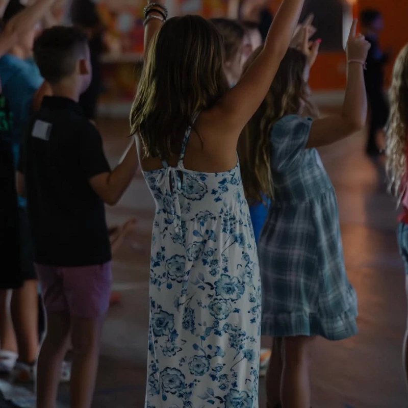 A group of children, seen from behind, raise their hands and point up towards a white cross on a colorful wall in a dimly lit room.