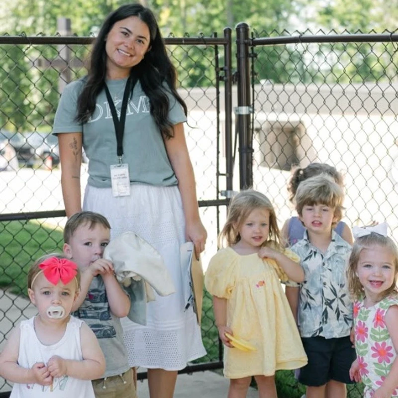 A smiling female teacher with long dark hair stands outdoors with five young children in front of a chain-link fence, posing for a group photo.