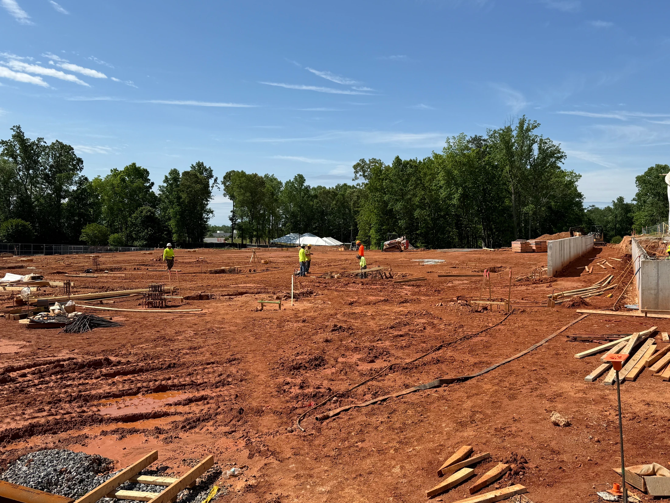 A wide view of a large construction site with red clay soil. Several workers in safety vests are present among building materials and foundation work.
