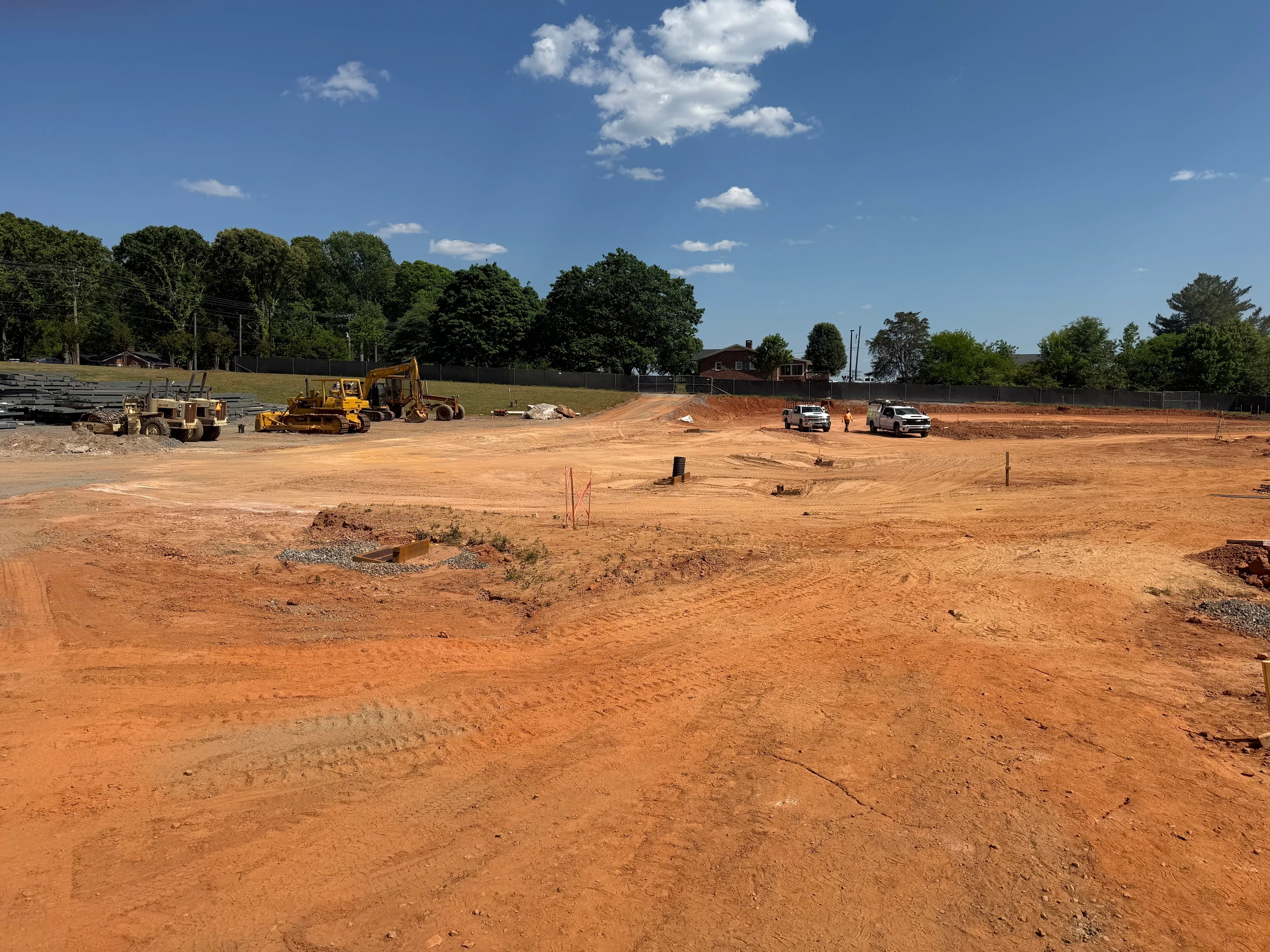A wide shot of a construction site with graded red clay earth. Heavy machinery, work trucks, and trees are in the background under a sunny blue sky.