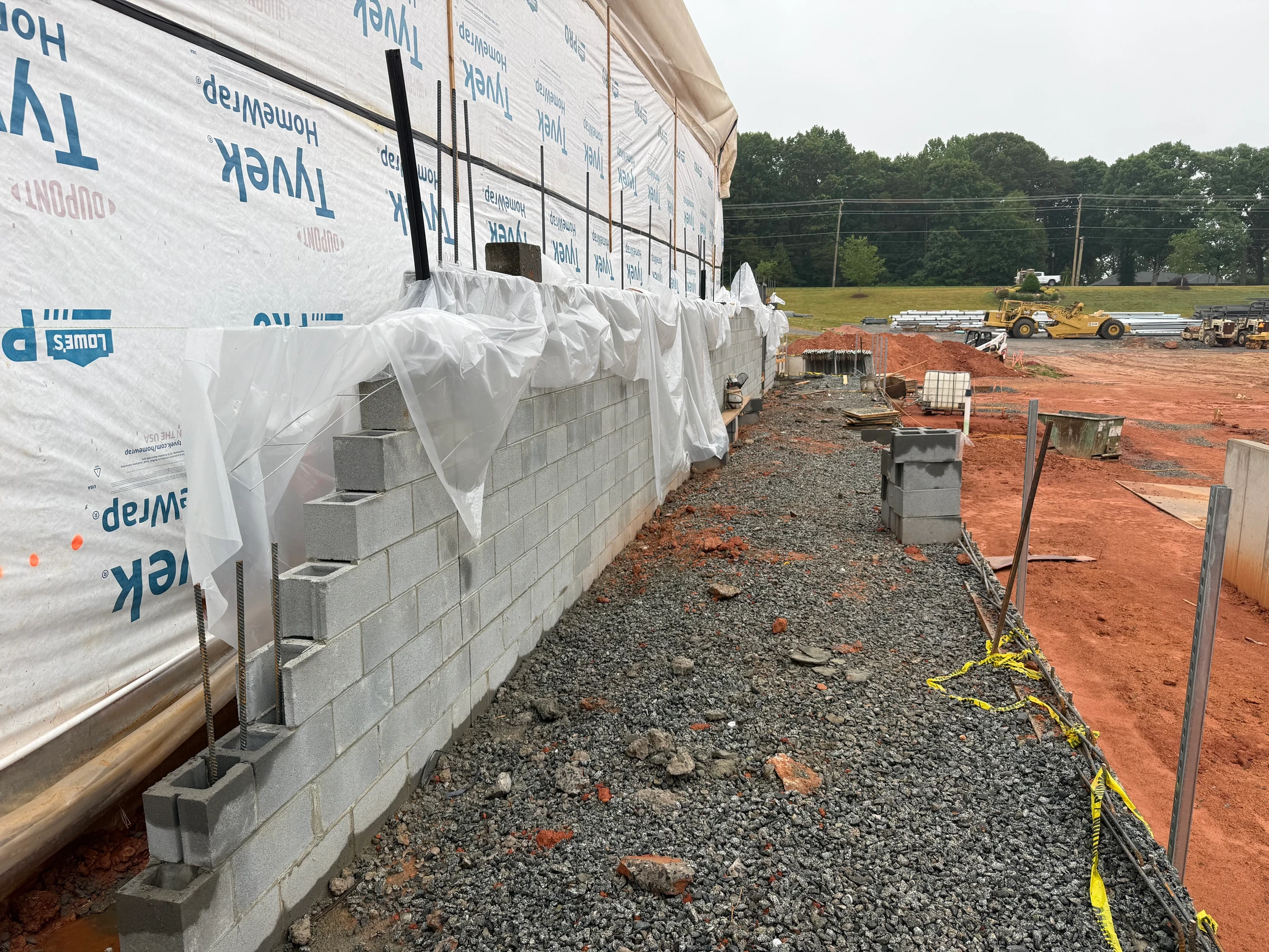 A new concrete block foundation wall at a construction site. The ground is gravel and red dirt, and the upper part of the building is covered in Tyvek wrap.