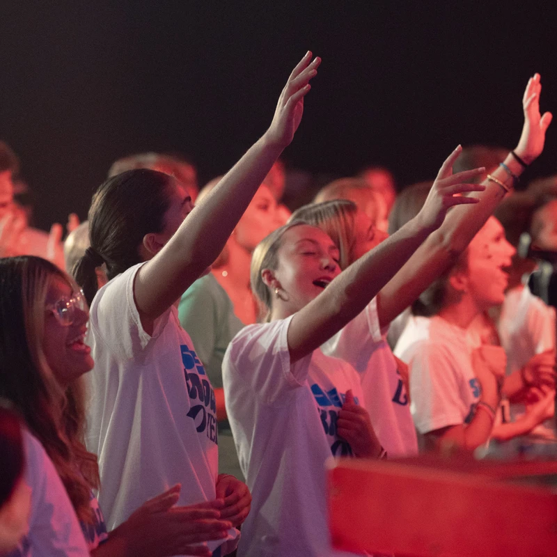 A group of young women in matching white t-shirts sing with their hands raised in a crowd, illuminated by pink and red stage lighting.