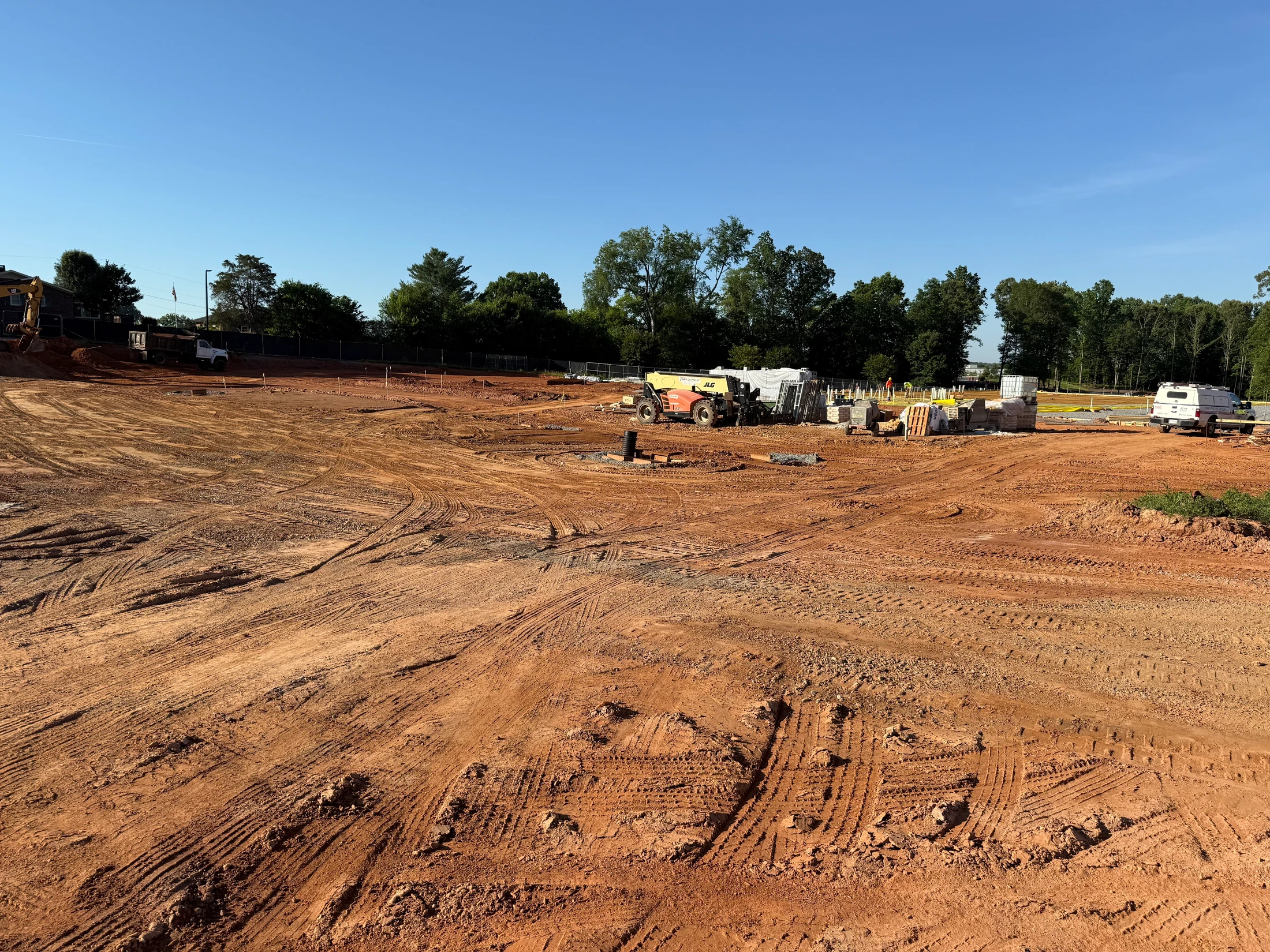 A wide view of a construction site with red clay ground covered in tire tracks. Equipment and building materials are in the background under a clear blue sky.