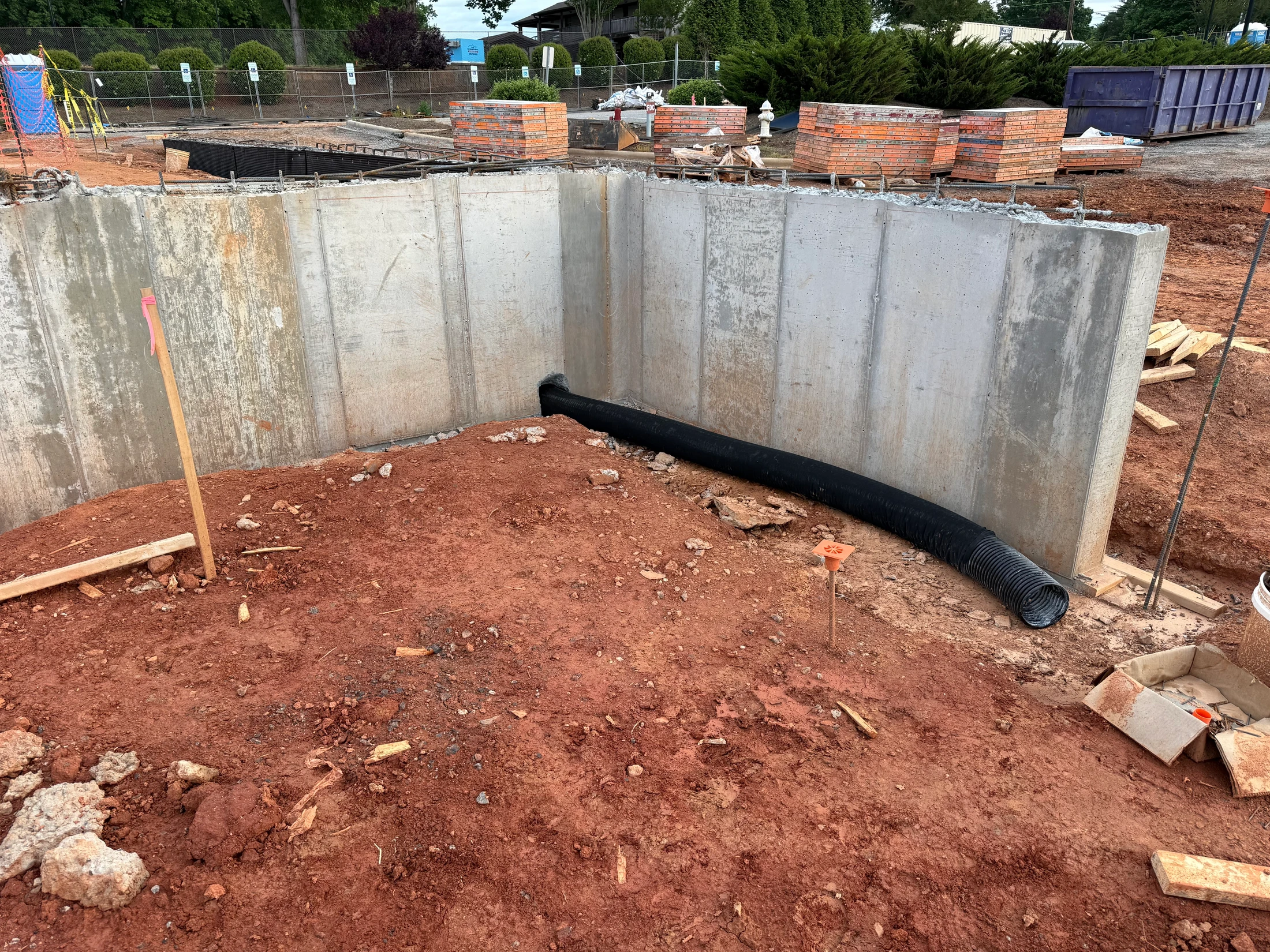 The corner of a new concrete foundation at a construction site, with red clay soil inside and a black corrugated drainage pipe running along the base.