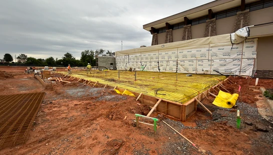 A construction site with workers preparing a large foundation with wooden forms, a yellow vapor barrier, and rebar next to a building under construction.