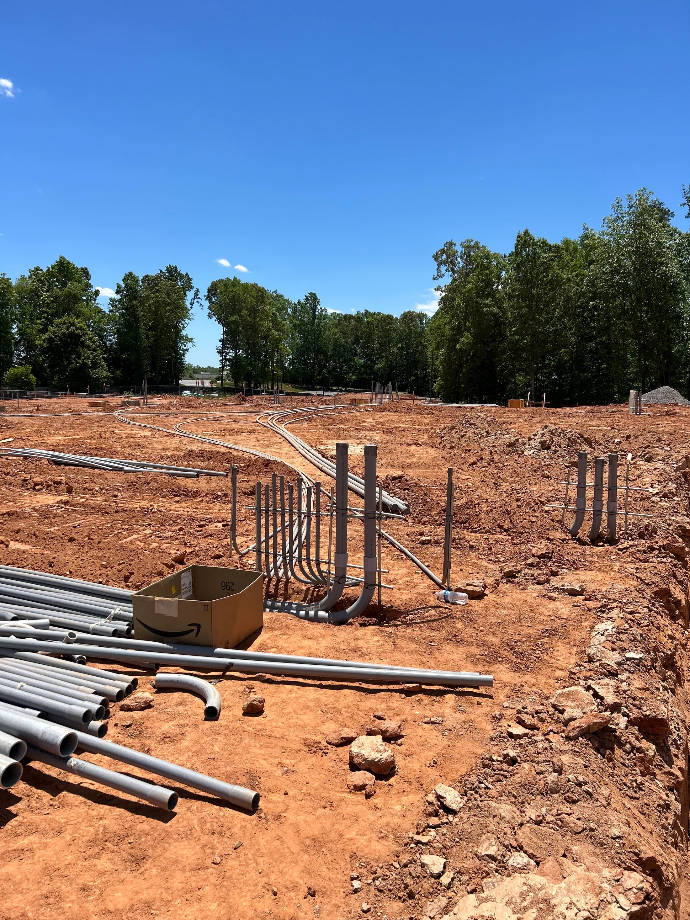 A construction site on a sunny day with red dirt ground. Grey electrical conduits and pipes are laid out in preparation for a new building's foundation.