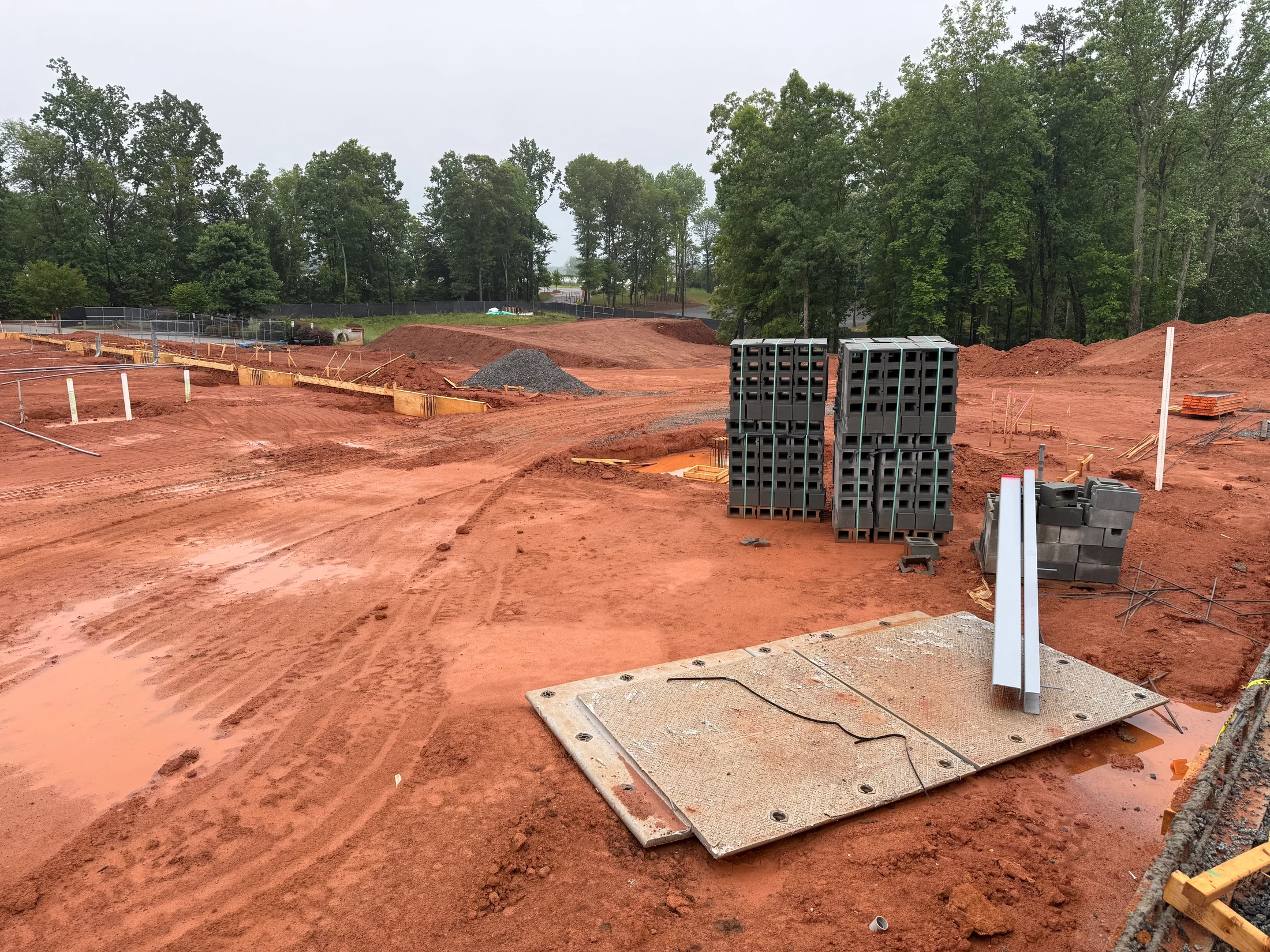 A wide view of a construction site with muddy red earth. Stacks of concrete blocks and other building materials sit near a partially built foundation.