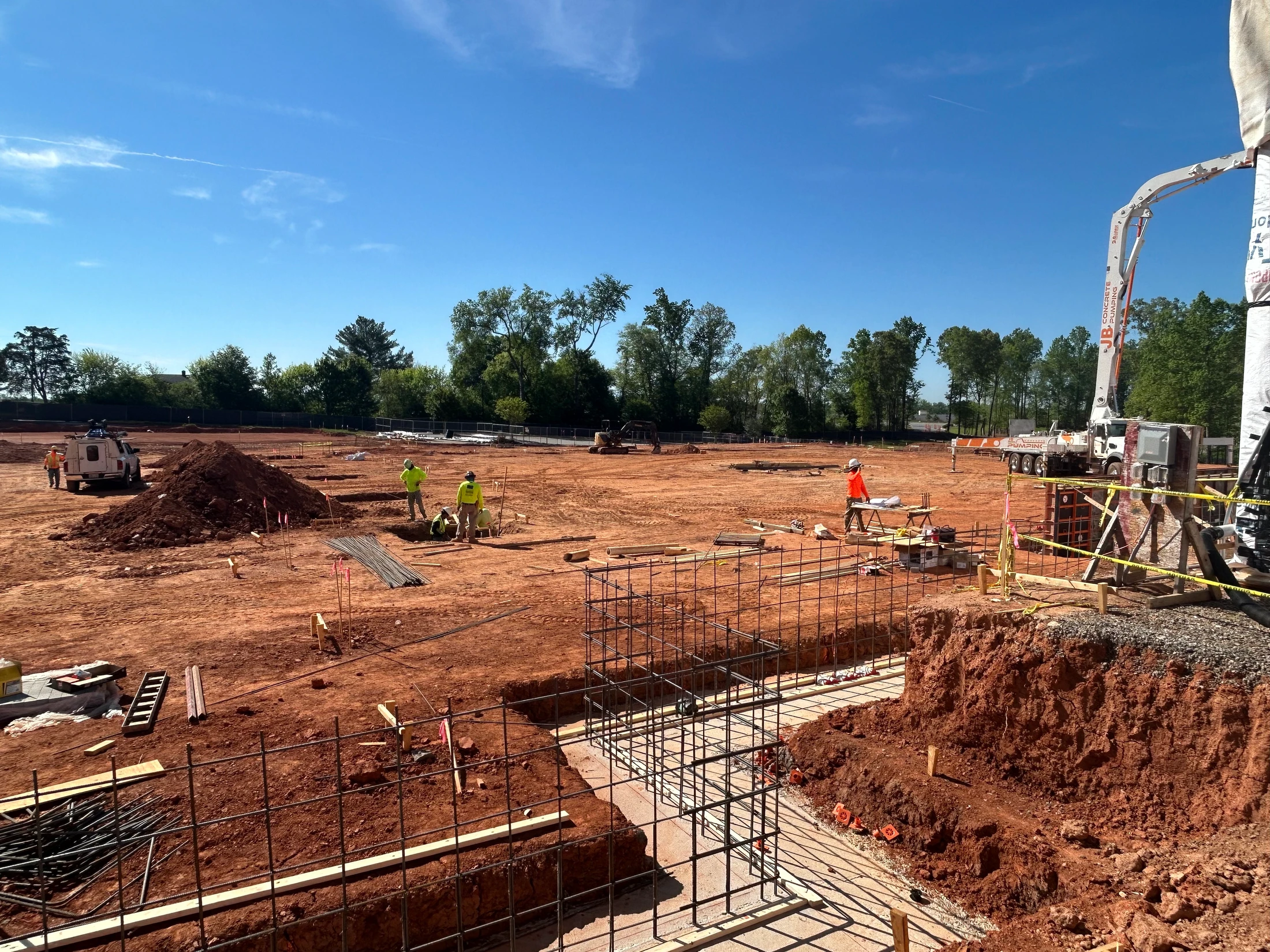 A wide shot of a construction site with red dirt under a clear blue sky. Workers prepare rebar cages in trenches for a building's foundation.