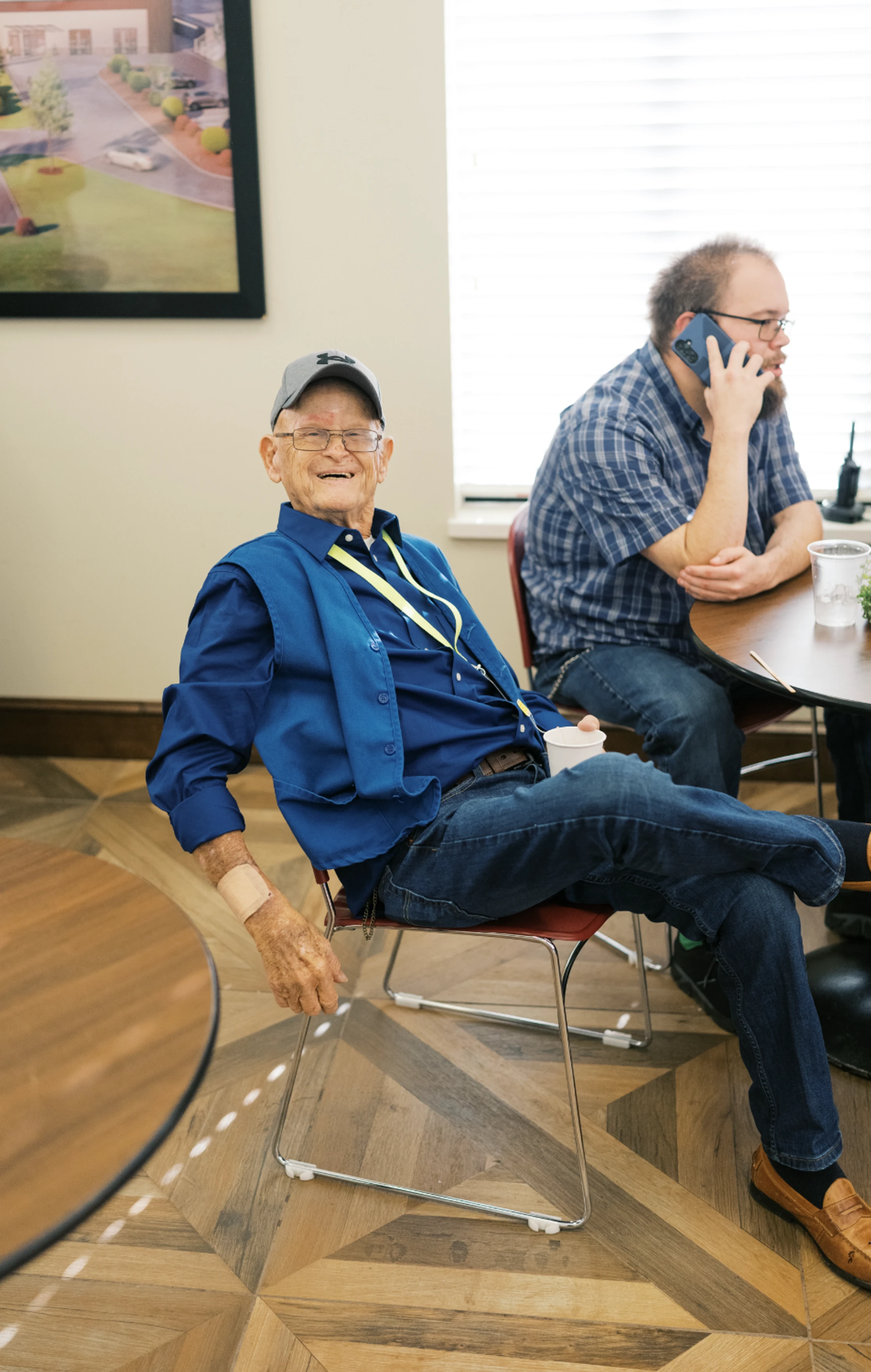 An elderly man in a blue shirt and baseball cap smiles while relaxing in a chair. In the background, another man is sitting at a table talking on the phone.