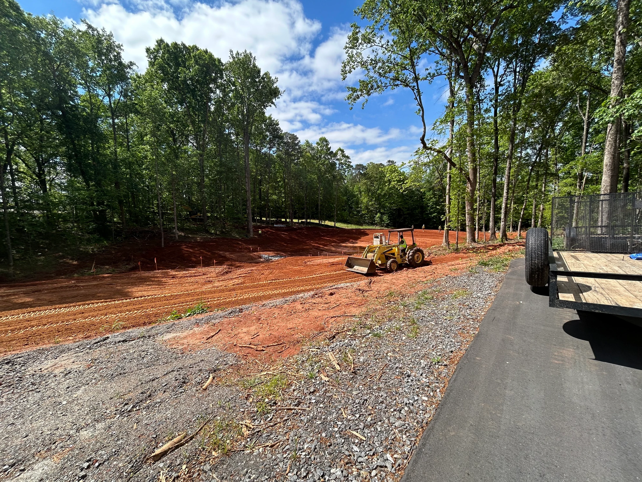 A yellow tractor grades a large patch of red clay at a construction site surrounded by a green forest under a blue sky with white clouds.