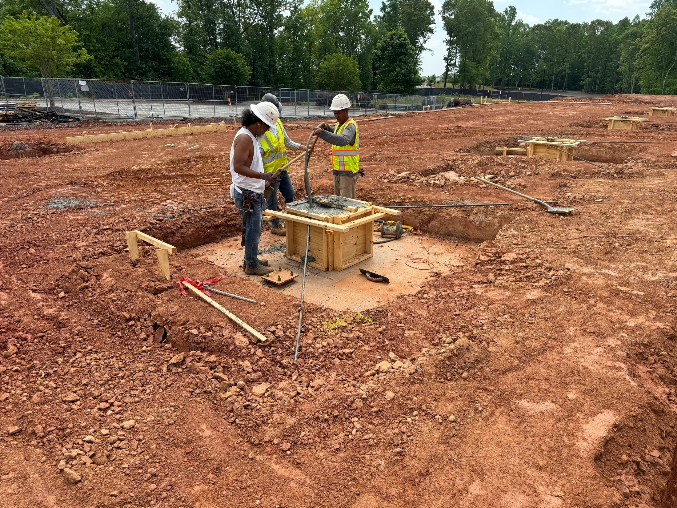 Three construction workers in hard hats and vests pour concrete into a wooden form on a red dirt construction site with other foundation footings nearby.