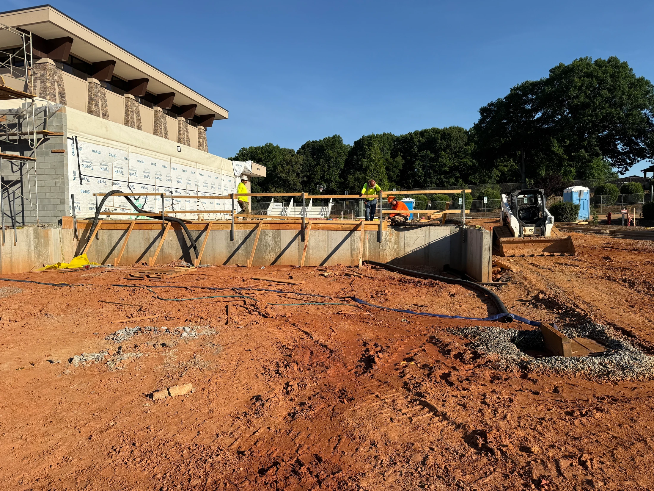 Construction workers on a concrete wall at a building site with red clay in the foreground, a skid steer, and trees in the background under a blue sky.