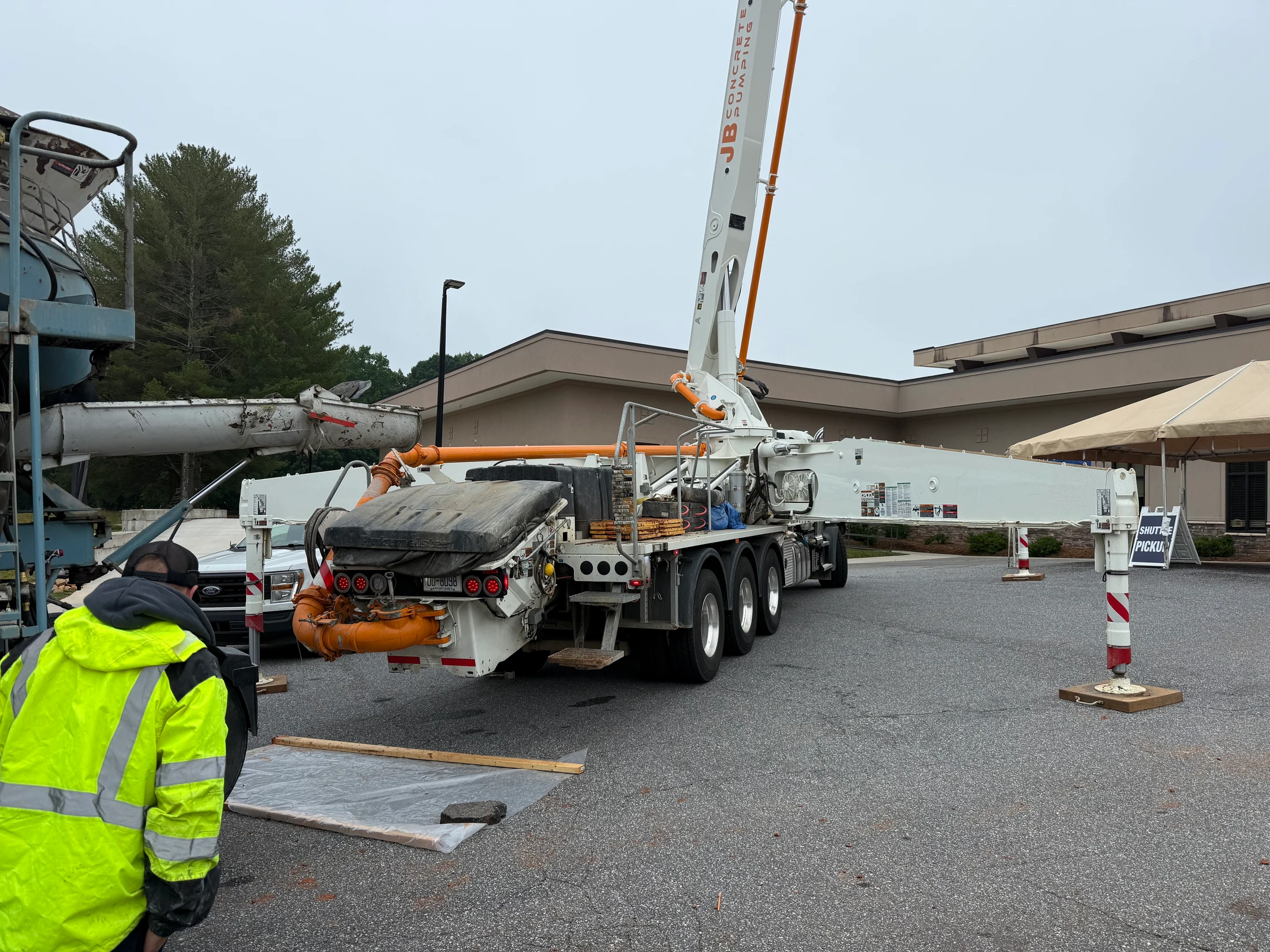 A worker in a yellow safety jacket stands next to a large, white concrete pump truck with an orange boom parked in a lot on an overcast day.