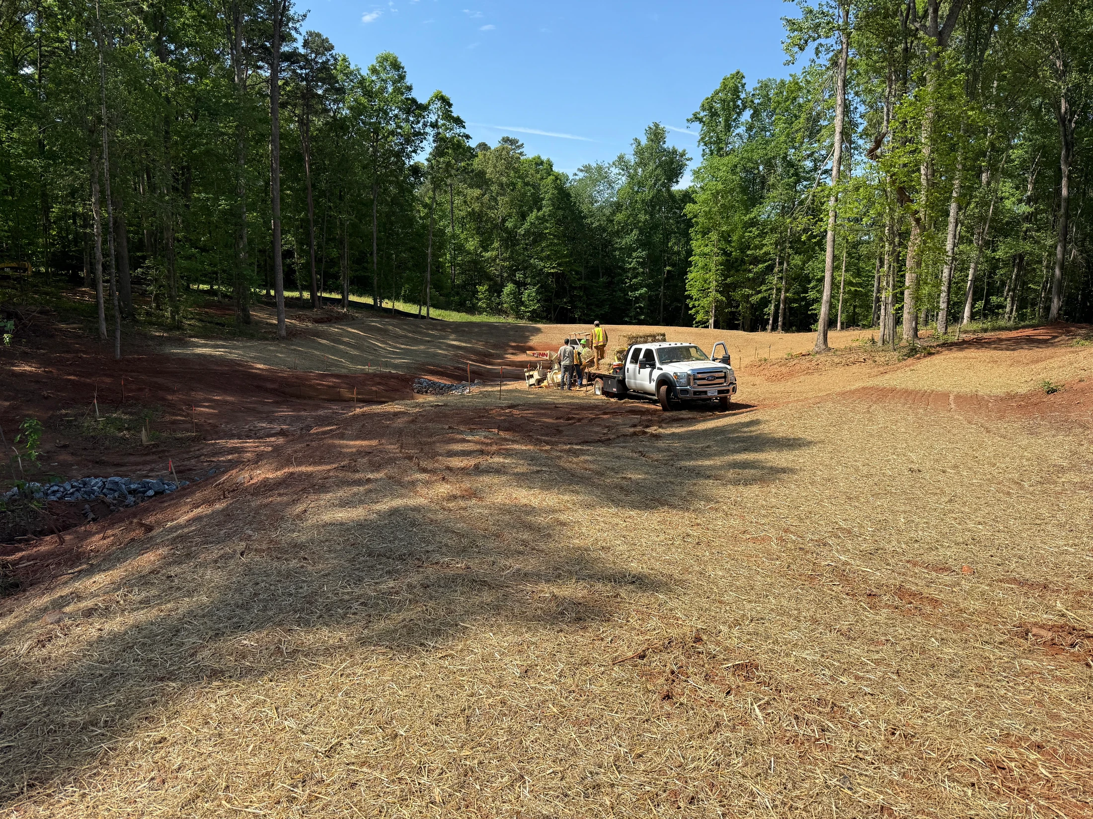 A crew of workers next to a white truck spreads straw over freshly graded red soil on a construction site, with a dense forest in the background on a sunny day.