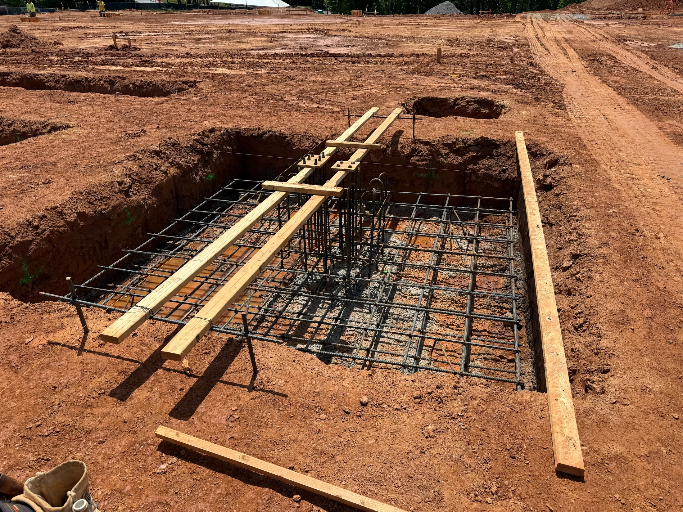 A square pit dug into red earth at a construction site, containing a steel rebar cage and wooden supports, prepared for a concrete foundation footing.