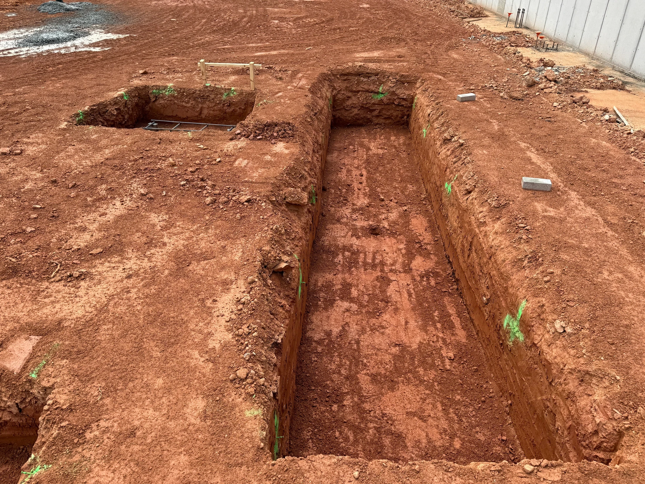 An overhead view of freshly dug foundation trenches in reddish-brown soil at a construction site, with green survey marks and a concrete wall in the background.