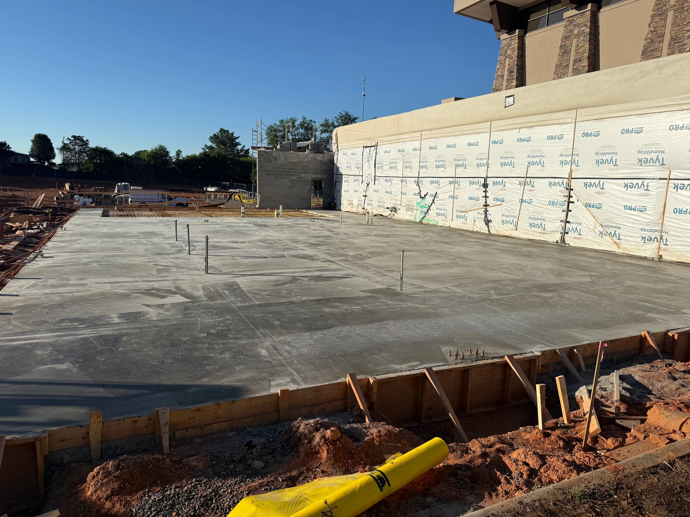 A large, freshly poured concrete slab at a construction site next to a building with walls covered in white Tyvek wrap under a clear blue sky.
