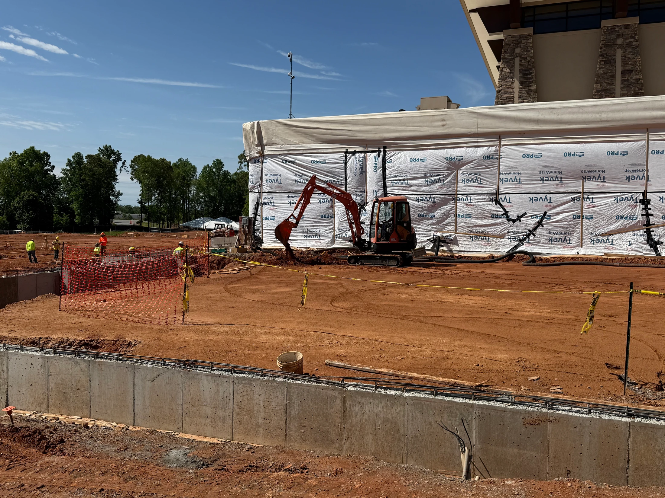 A sunny construction site shows a mini-excavator by a wrapped building, workers on red soil, and a new concrete foundation wall with rebar.