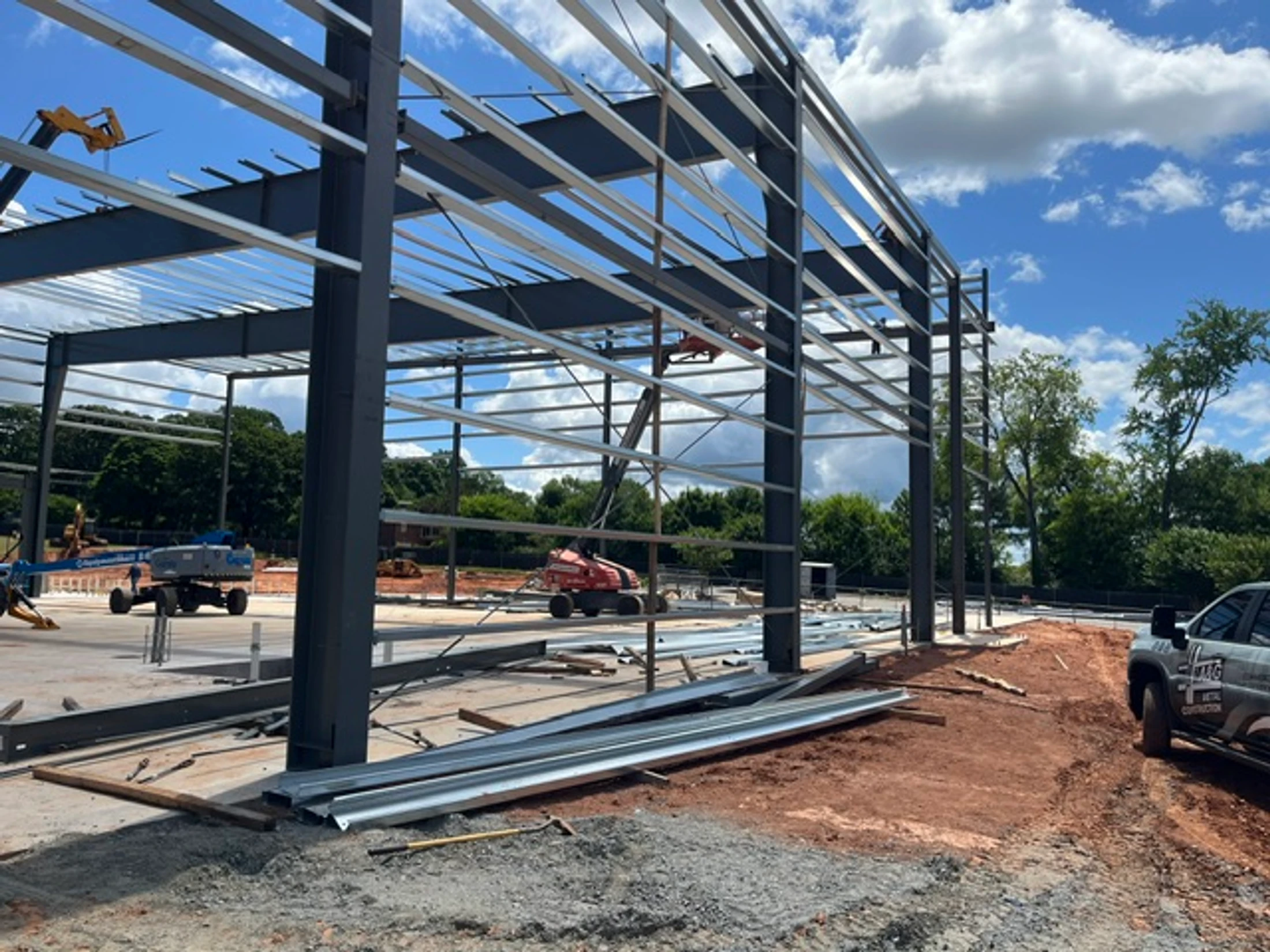 The steel framework of a large building is being erected on a concrete slab, with construction equipment visible under a blue sky with clouds.