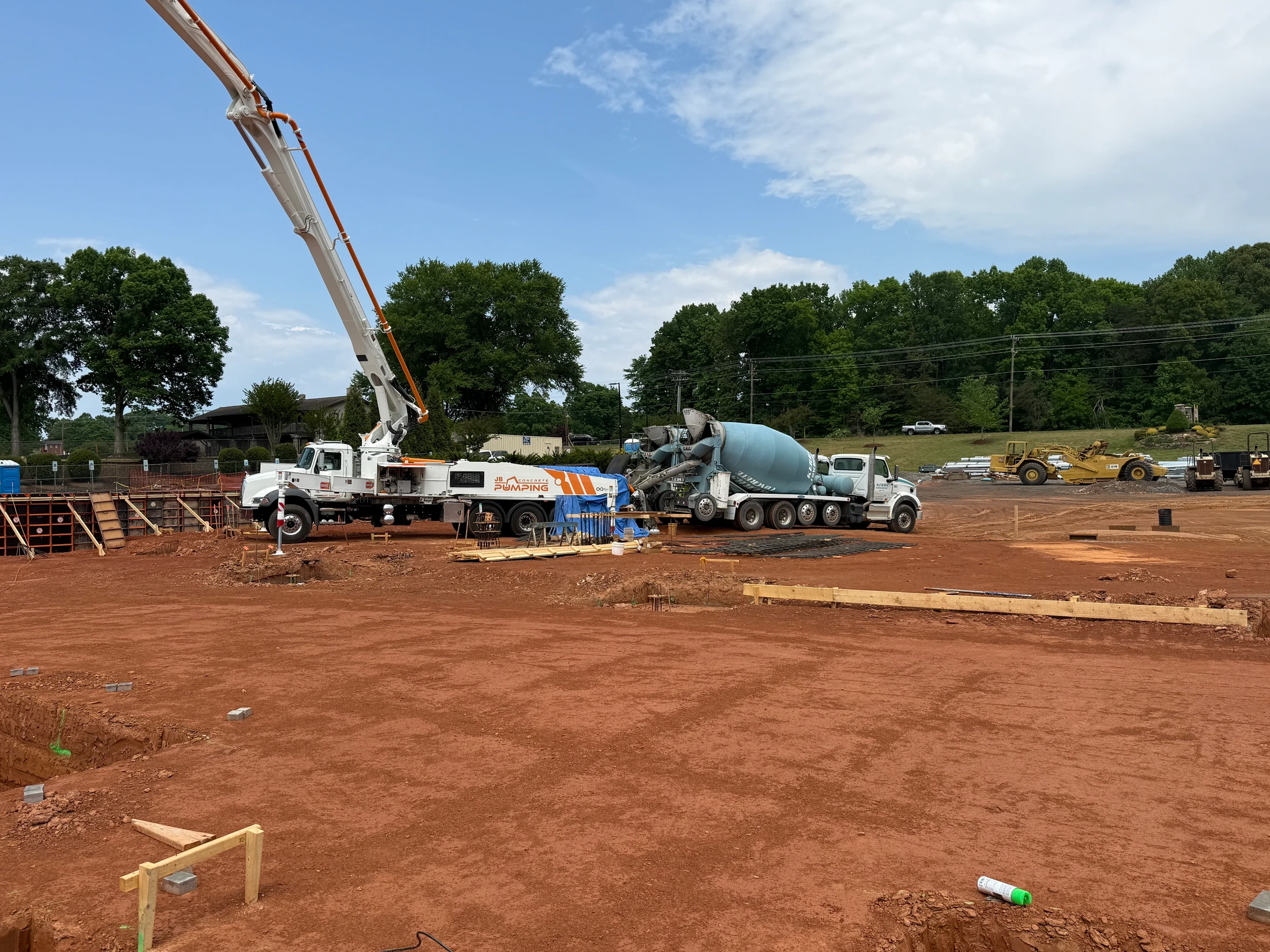 A large construction site with red clay soil, featuring a concrete pump truck with an extended boom and a cement mixer truck ready to pour foundations.