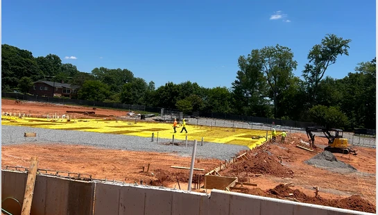 A construction site on a sunny day, with a concrete foundation wall in the foreground and workers laying a yellow vapor barrier on a gravel bed.