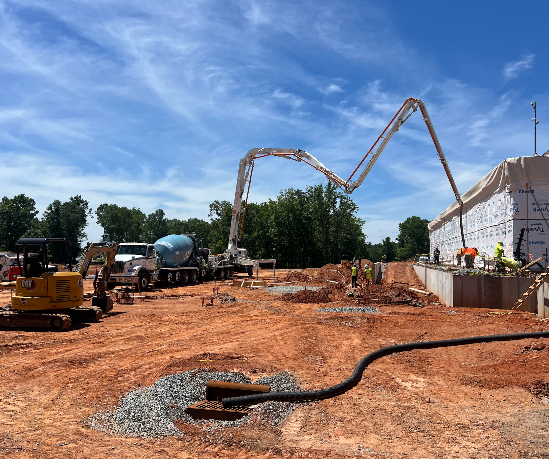 A wide view of a construction site with a concrete pump truck extending its long boom to pour a foundation, with other heavy machinery and workers nearby.