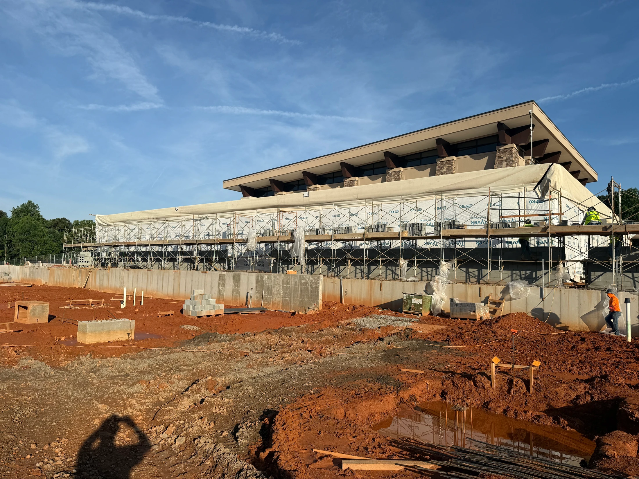 A wide shot of a construction site with a modern building being built. Scaffolding covers the lower level, and the foreground is red clay mud.