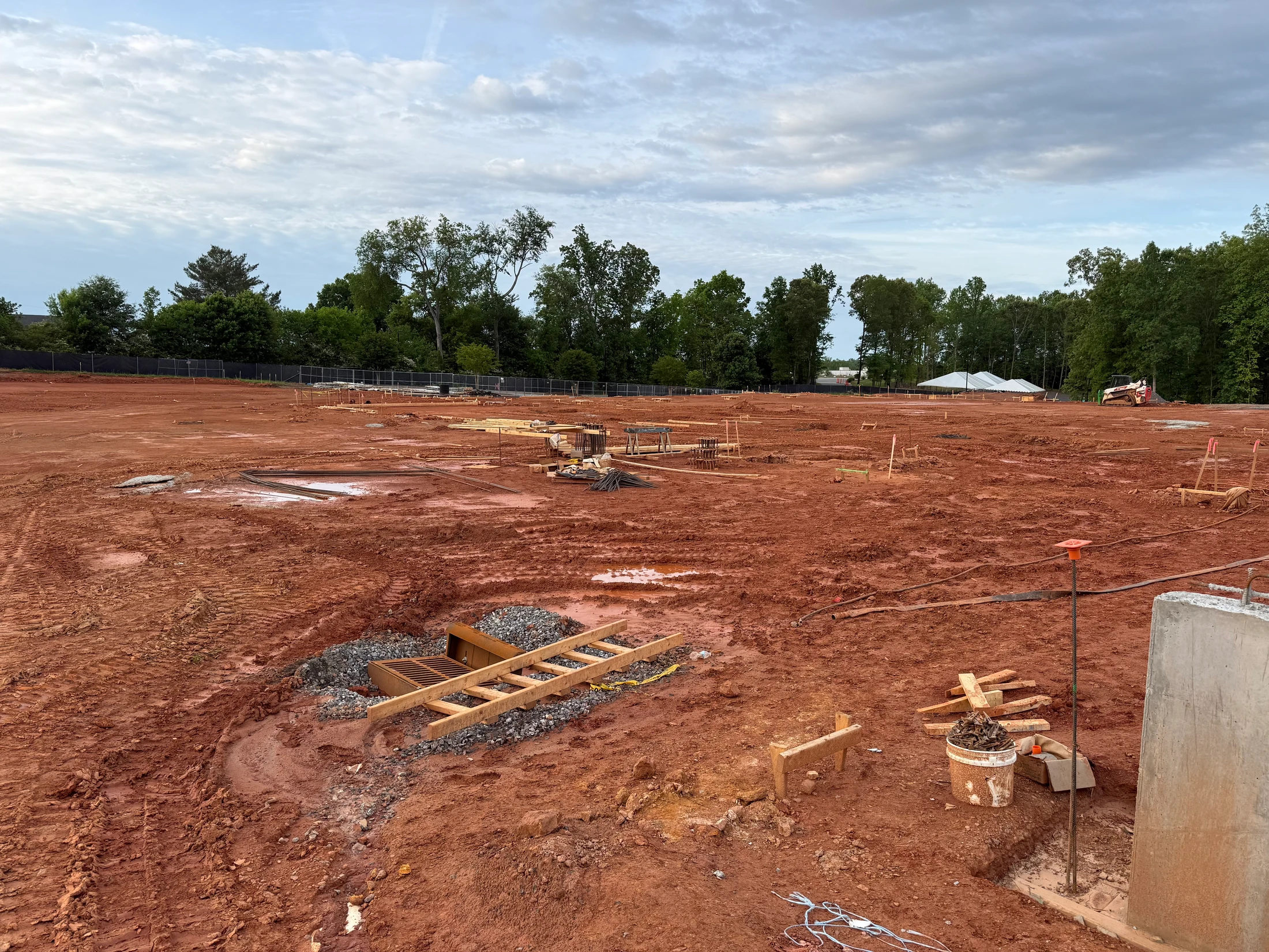 A wide shot of a construction site with muddy red clay ground. Building materials like wood and rebar are scattered about. Trees line the background.