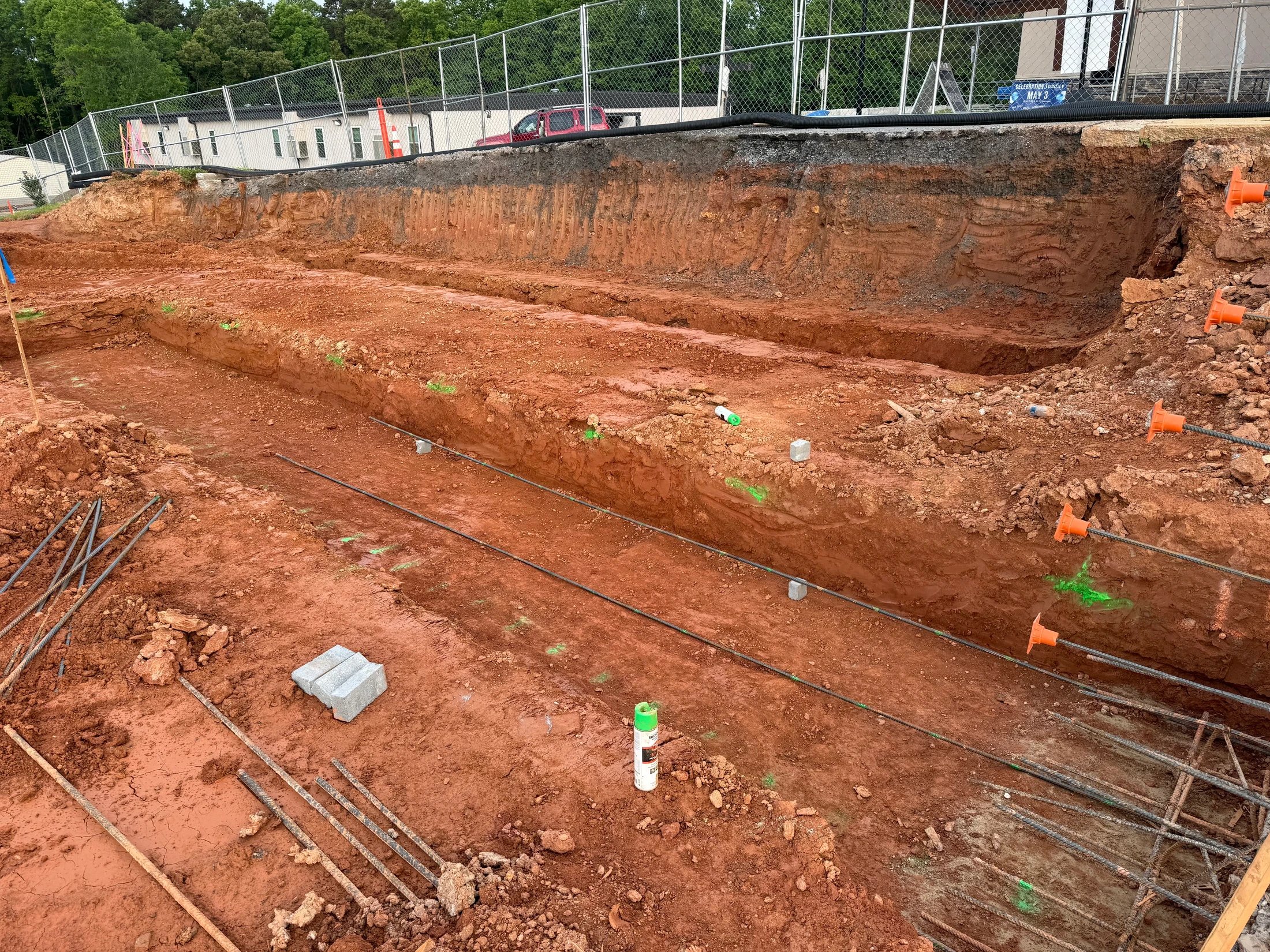 A construction site with excavated red earth where long trenches are dug for a foundation. Steel rebar is laid out in the trenches, preparing for concrete.