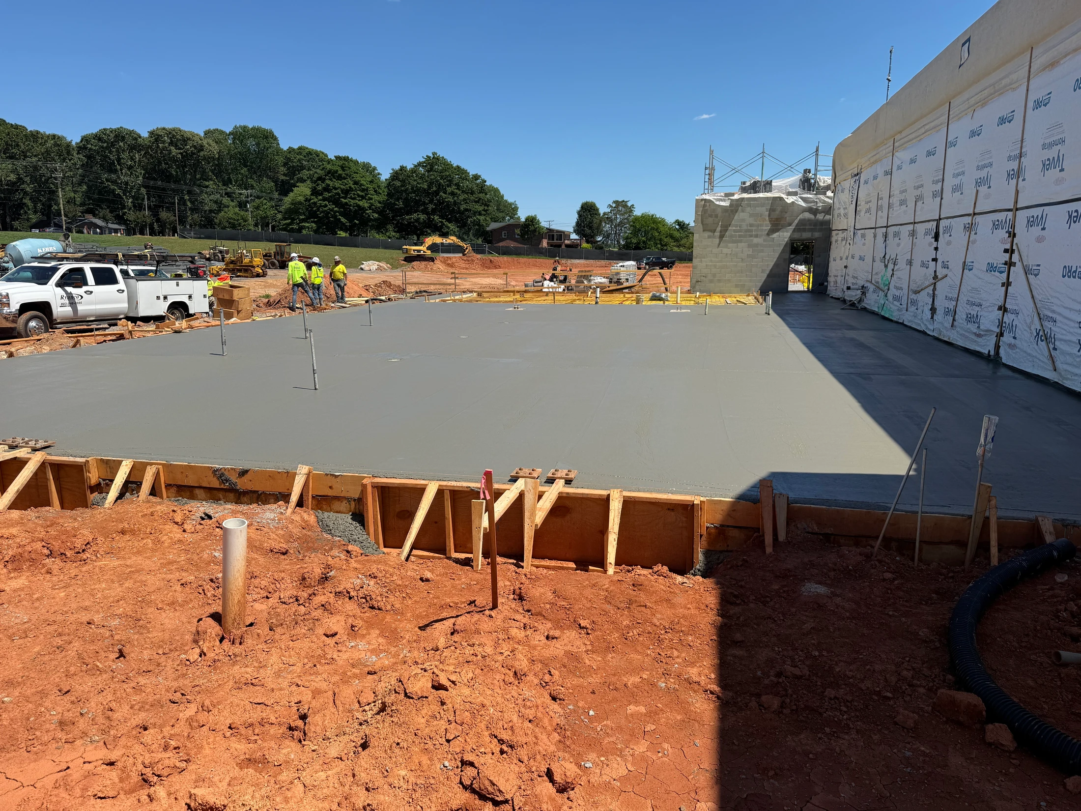A large, smooth slab of freshly poured wet concrete at a construction site, with wooden formwork in the foreground and a partially built wall to the right.