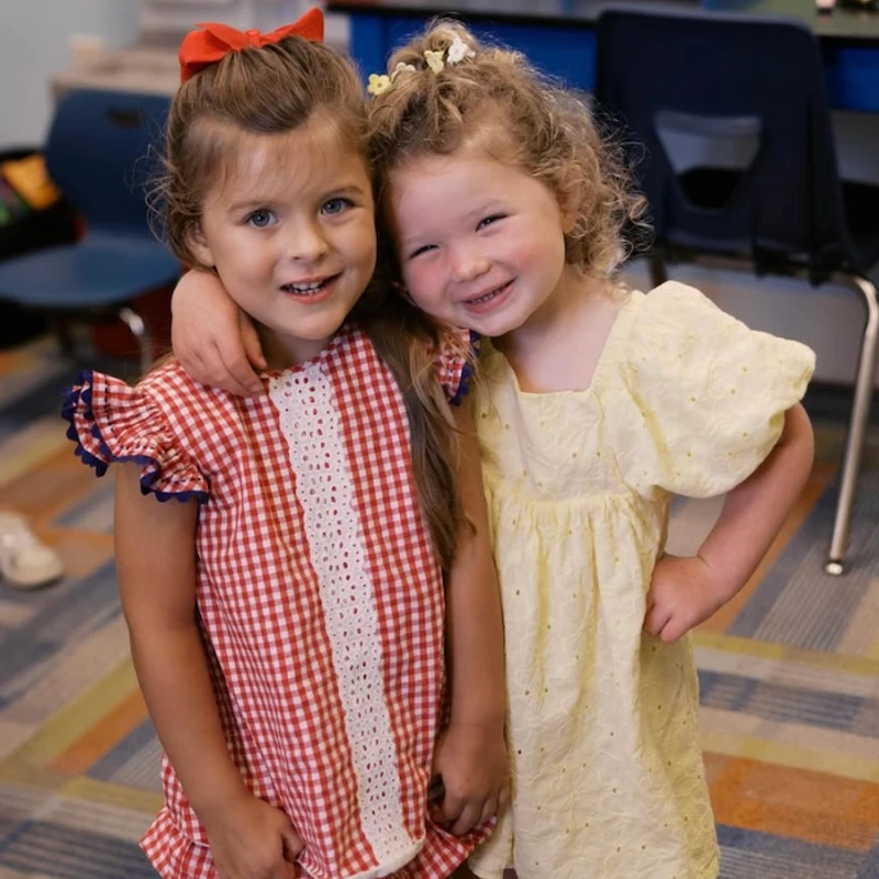 Two smiling young girls embrace in a classroom. One wears a red gingham outfit with a red bow, and the other wears a yellow dress with flower clips in her hair.