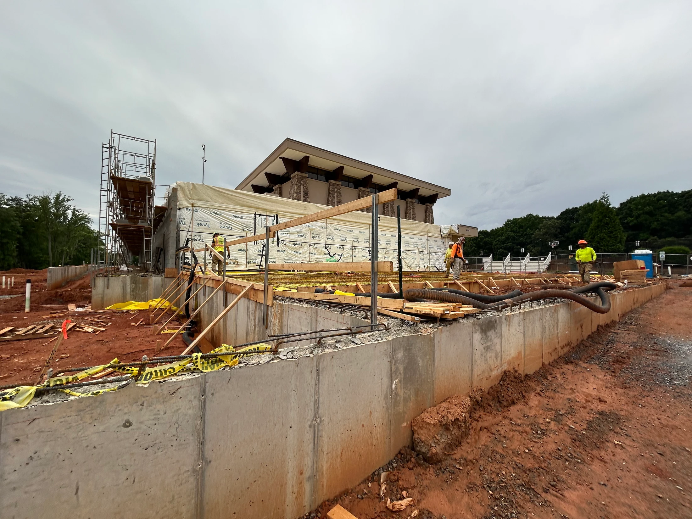 A construction site with workers standing on a wooden frame over a concrete foundation. A partially built building with scaffolding is in the background.