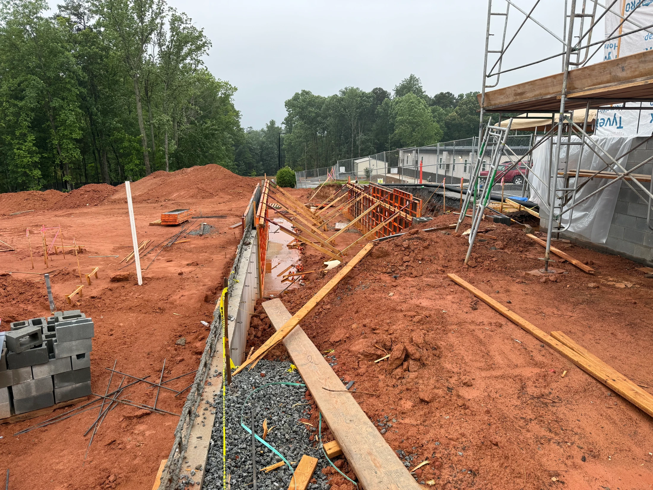 A construction site with red clay soil where a concrete retaining wall is being built using orange forms and wood braces. Scaffolding stands to the right.