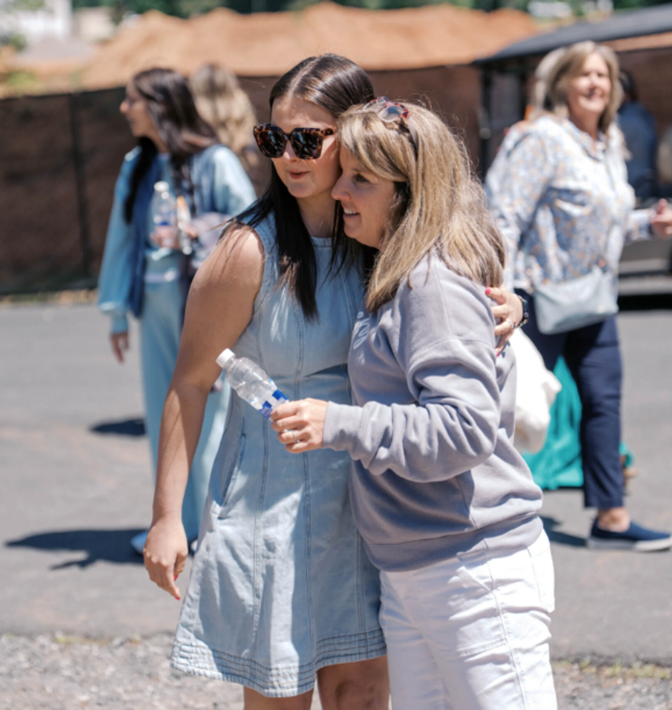 A young woman in a denim dress and sunglasses hugs an older woman in a grey sweatshirt. They are standing outdoors together on a sunny day.