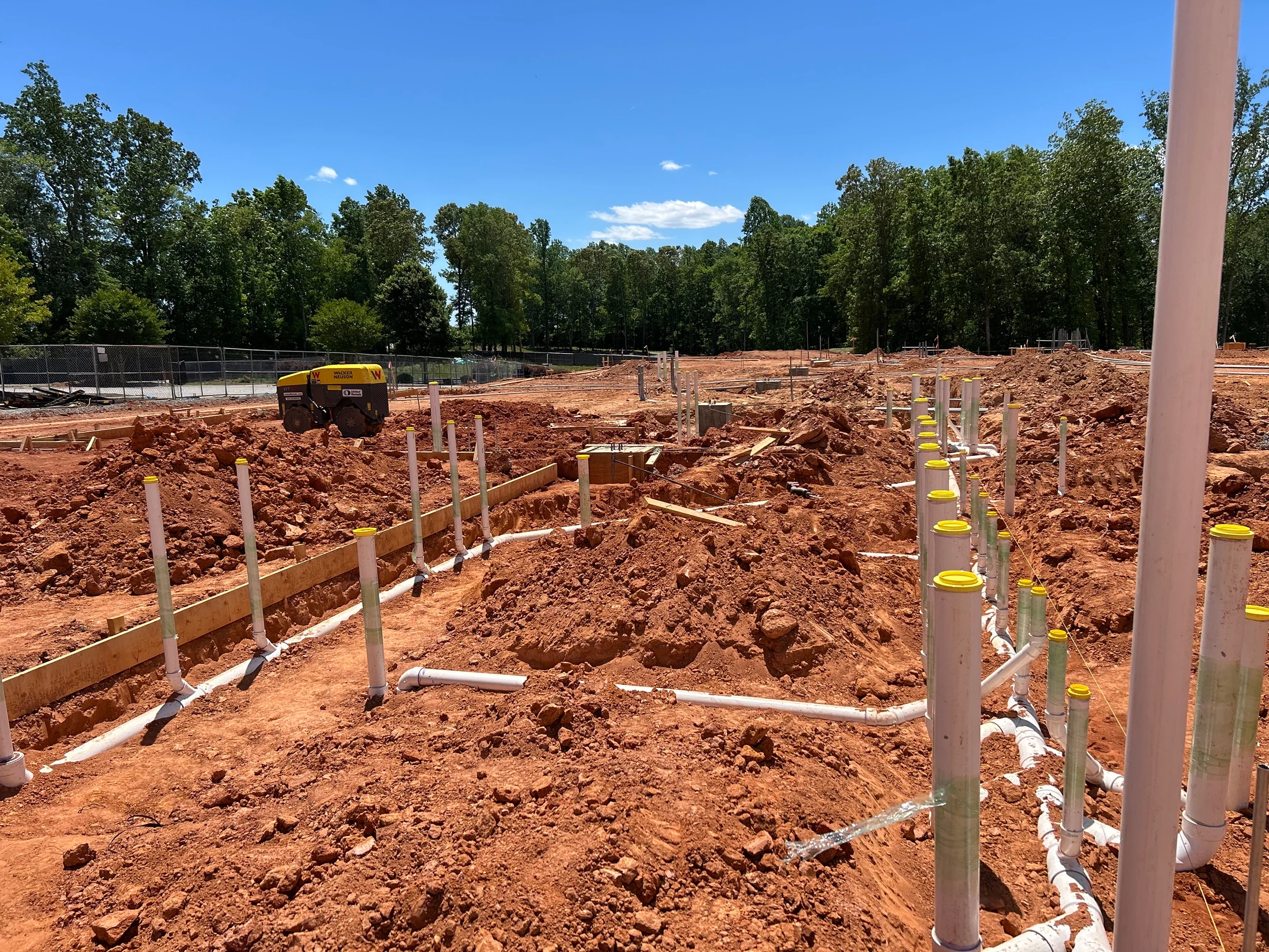 A construction site on red soil with white PVC pipes laid out for underground plumbing. Wooden foundation forms and a yellow compactor are also visible.
